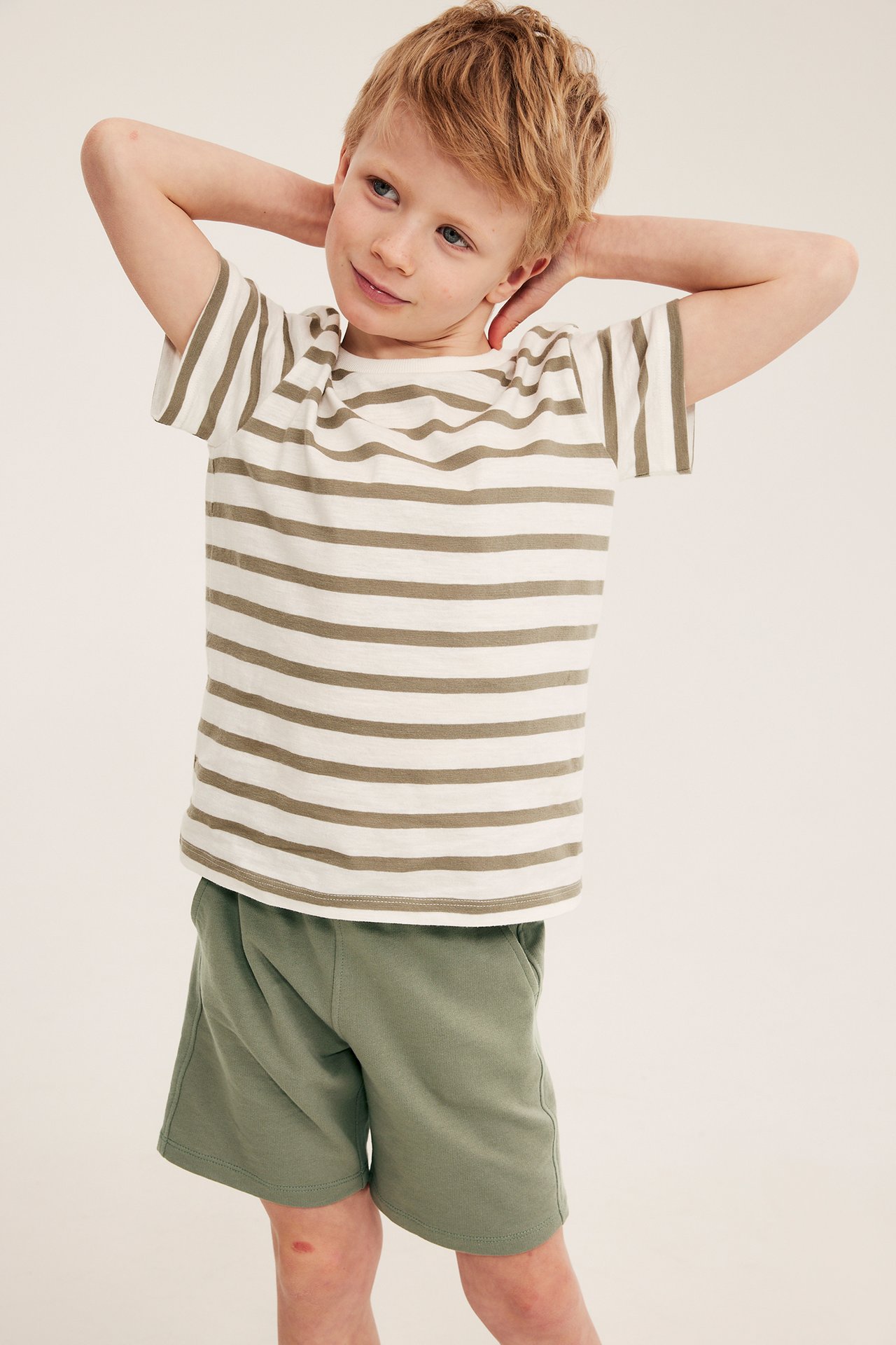 Edestäpäin kuvassa lapsi tummanvihreissä puuvillajersey-shortseissa ja valko-beigeraitaisessa t-paidassa.