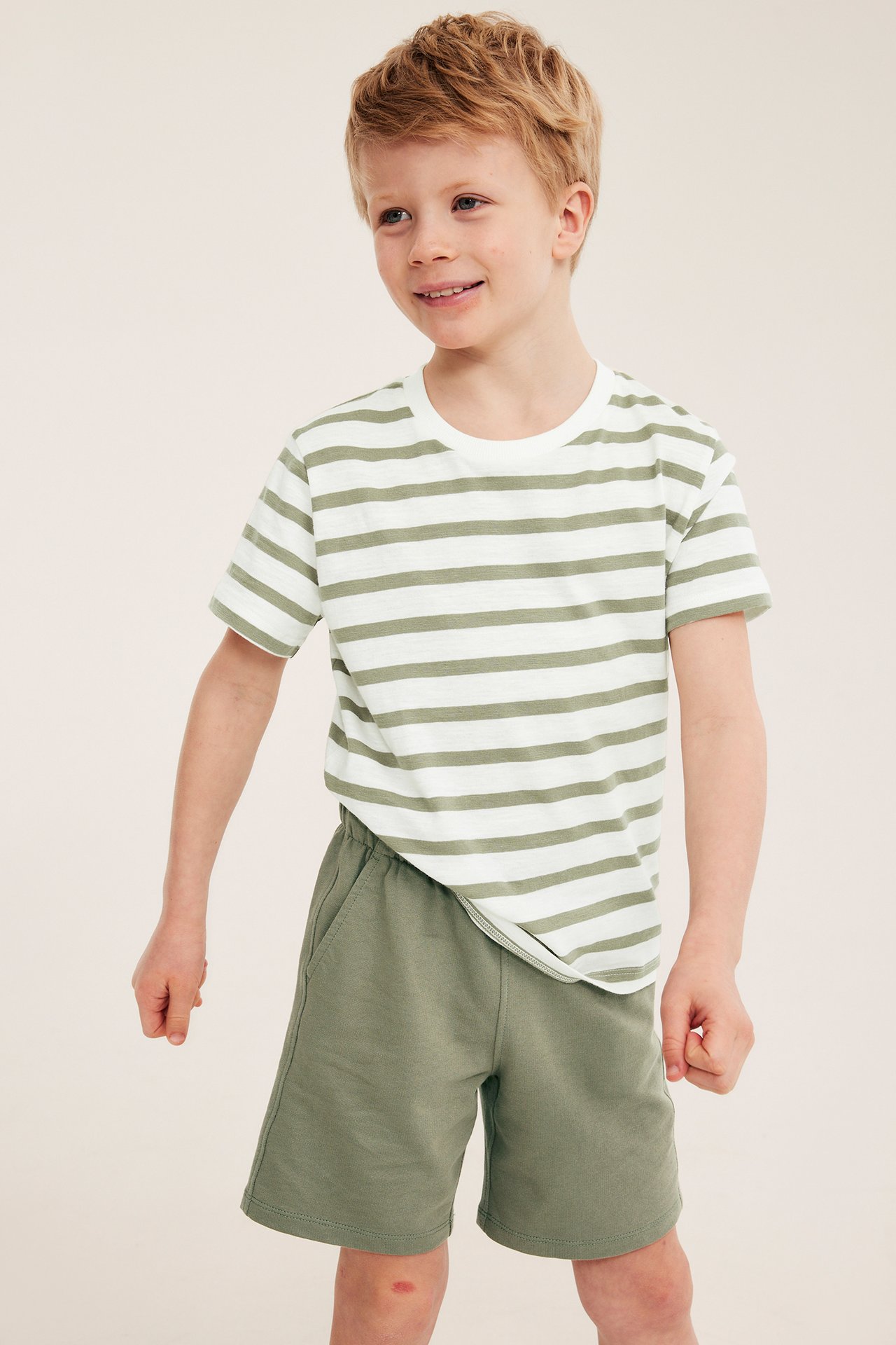 Framifrån, ett barn bär mörkgröna bomullsshorts och en vit- och ljusgrönrandig t-shirt.