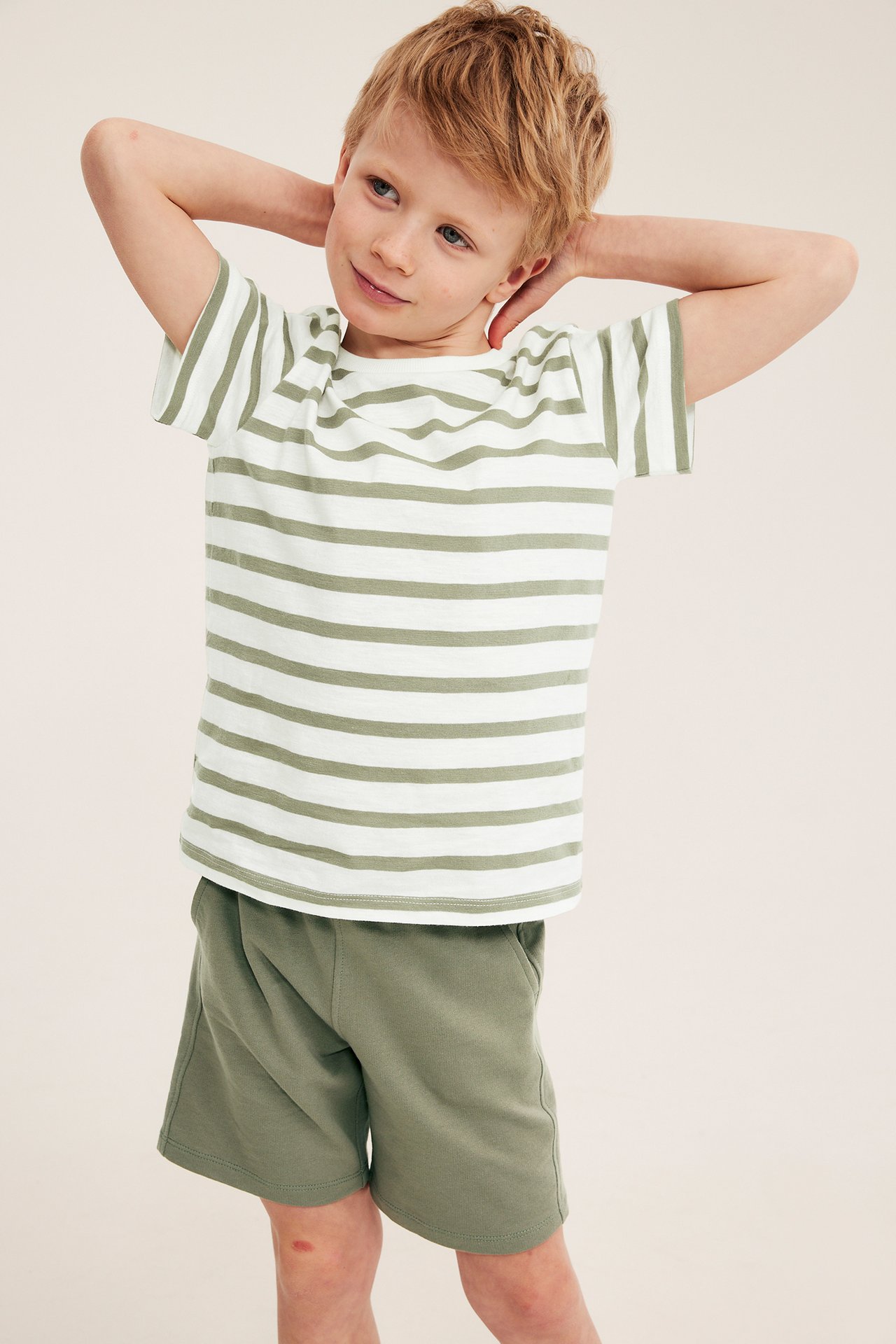 Edestäpäin kuvassa lapsi tummanvihreissä puuvillaisissa college-shortseissa ja valko-vaaleanvihreäraitaisessa t-paidassa.