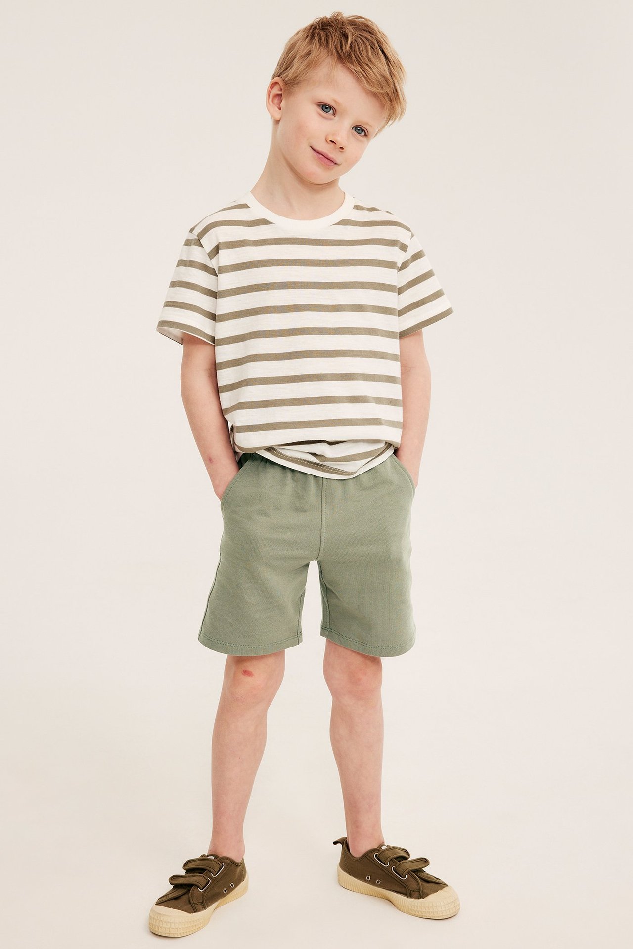Framifrån, ett barn i mörkgröna mjukisshorts i bomull, en randig t-shirt och olivgröna skor.