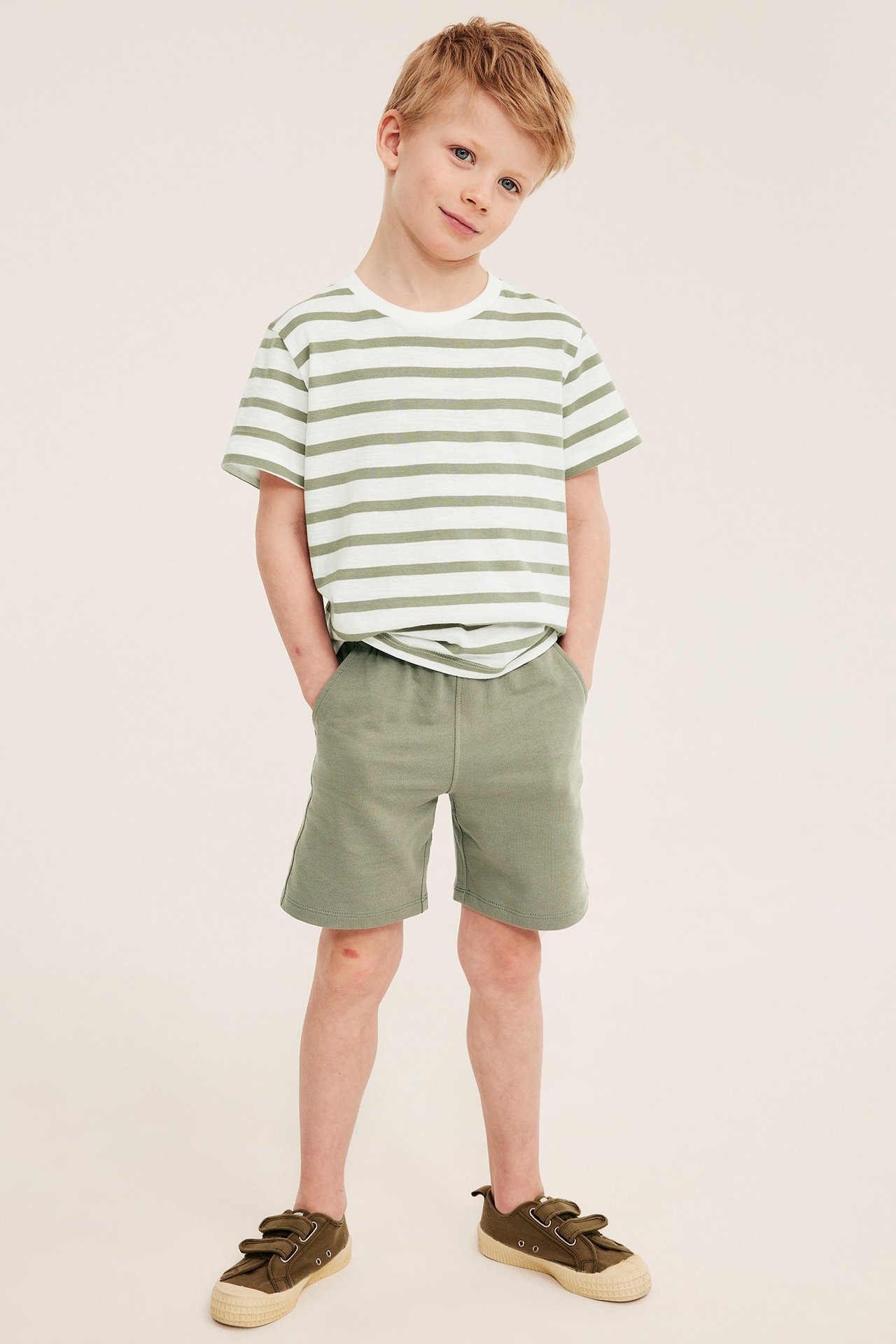 Etukuva: lapsi pitää tummanvihreitä puuvillajerseyshortseja, valko-vihreää raidallista t-paitaa ja tummanvihreitä kangaskenkiä.