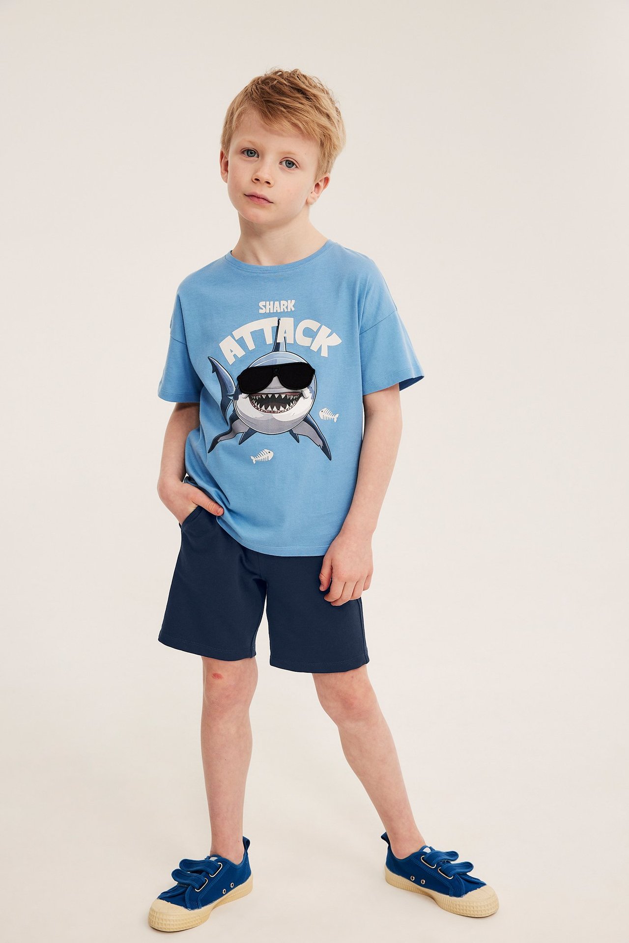 Barnets framifrån: mörkblå bomullsshorts, ljusblå t-shirt med hajtryck, och blå kardborresneakers.