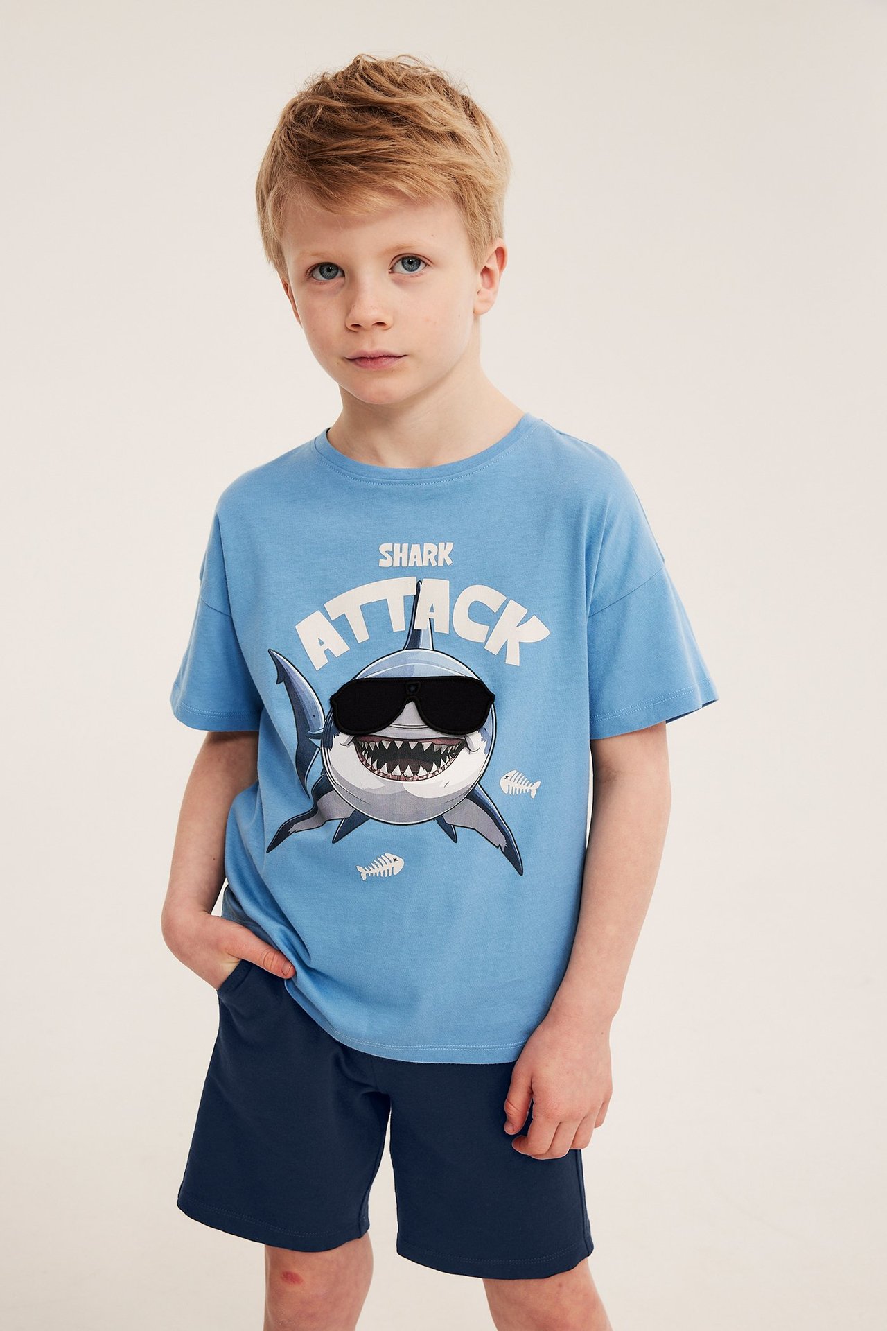 Framifrån, en liten pojke i ljusblå t-shirt med hajtryck och mörkblå enfärgade sweatshorts.