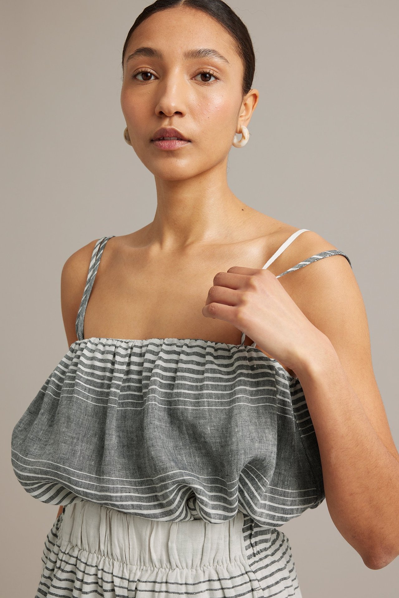 Frontvy: Kvinna i mörkgrått randigt linne, håller axelband. Bär matchande underdel och vita ringörhängen.