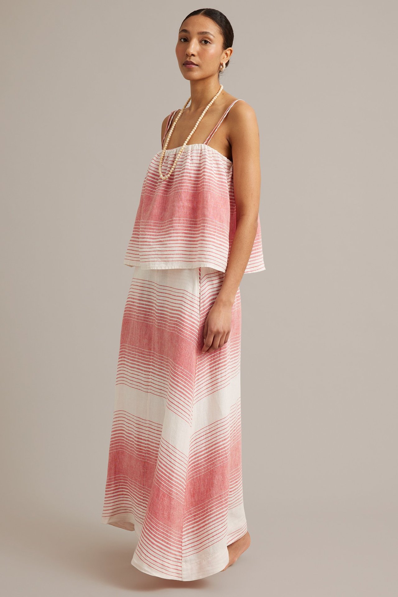 Kvinna i röd/vitrandigt linne av linne med smala axelband, matchande maxikjol och pärlhalsband, sidovy.