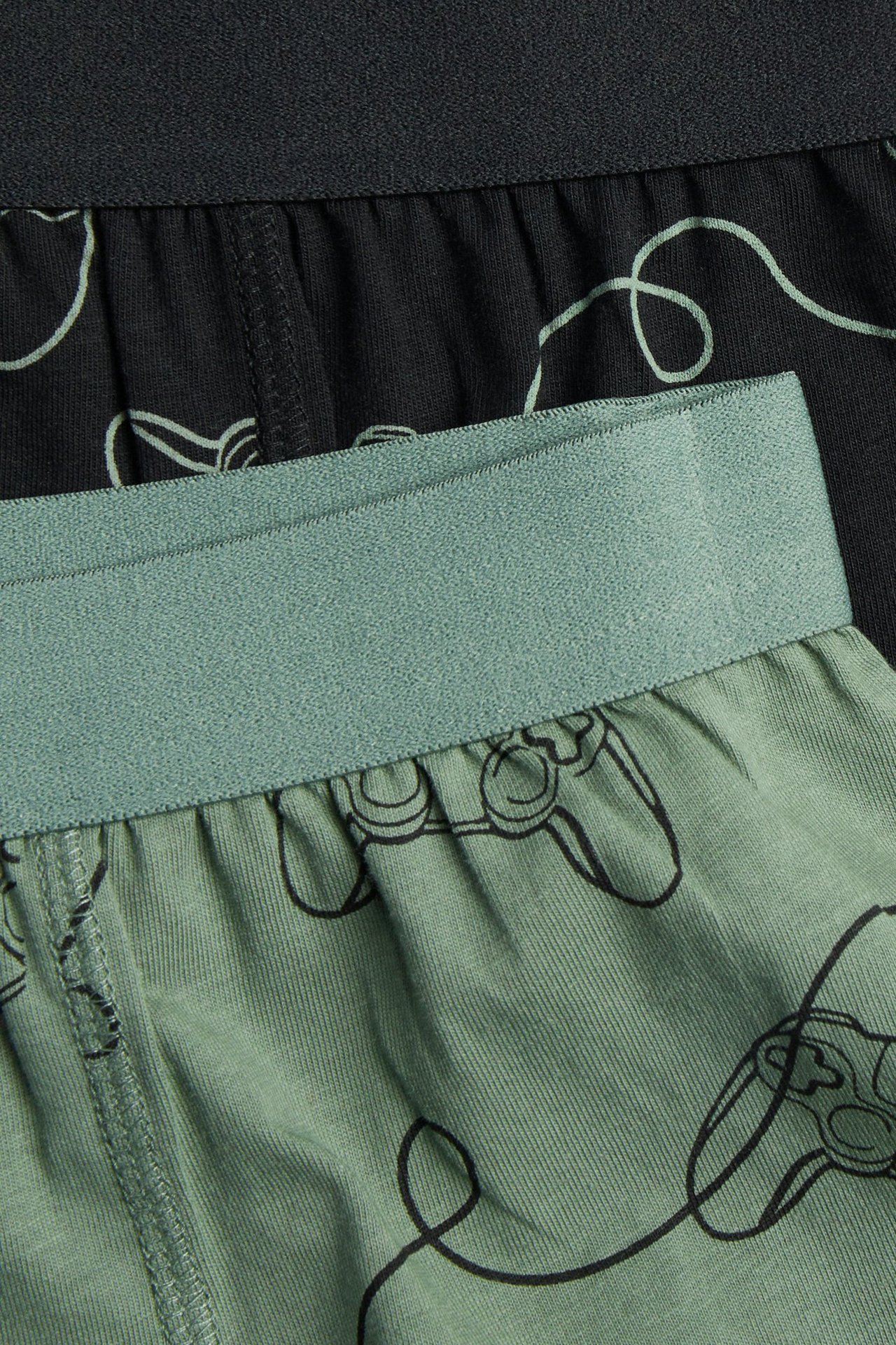 Närbild av två par boxerkalsonger för barn: en grön med spelkontroller och en svart med spellinjer.