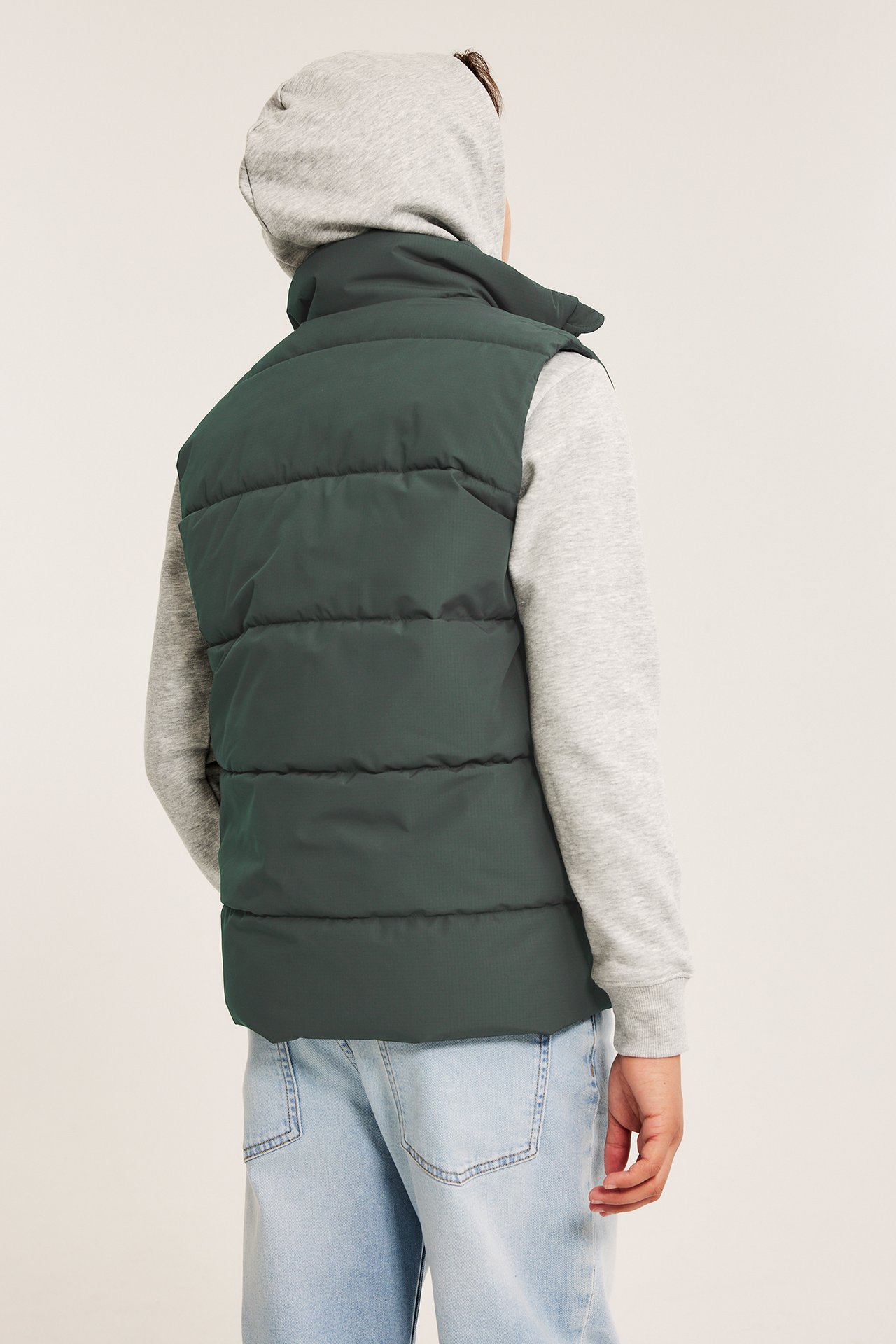 Widok od tyłu: dziecko w ciemnozielonej kamizelce puchowej na szarej bluzie z kapturem i jasnoniebieskich jeansach.