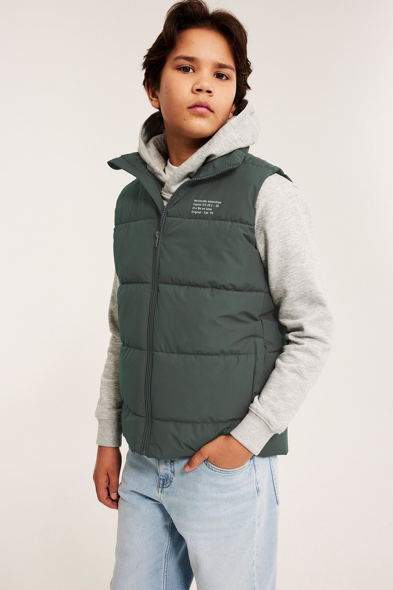 Widok z przodu: dziecko w ciemnozielonej kamizelce puchowej na szarej bluzie z kapturem i jasnoniebieskich jeansach.
