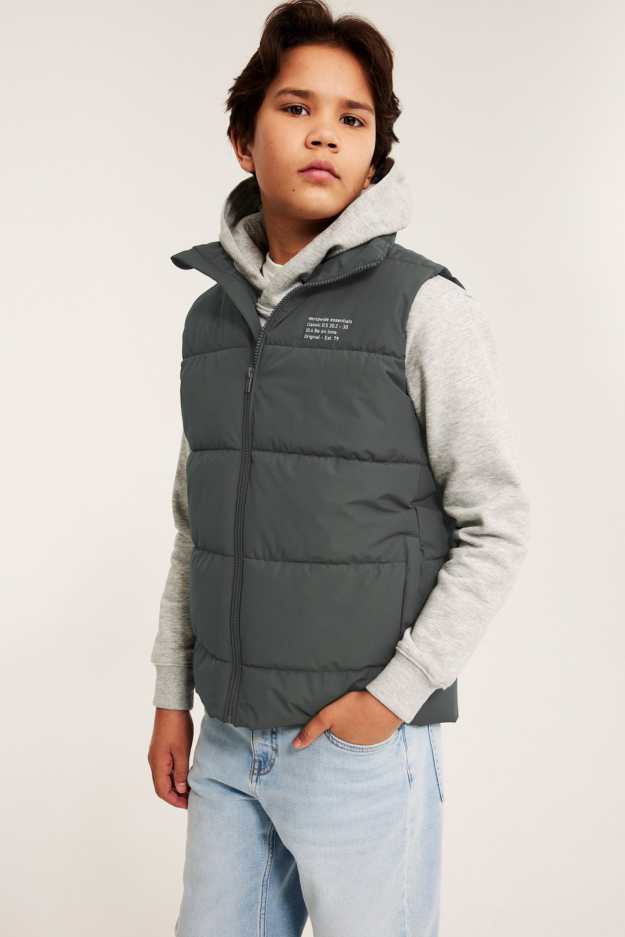 Dziecko z przodu w ciemnoszarej pikowanej kamizelce, jasnoszarej bluzie z kapturem i jasnoniebieskich dżinsach.