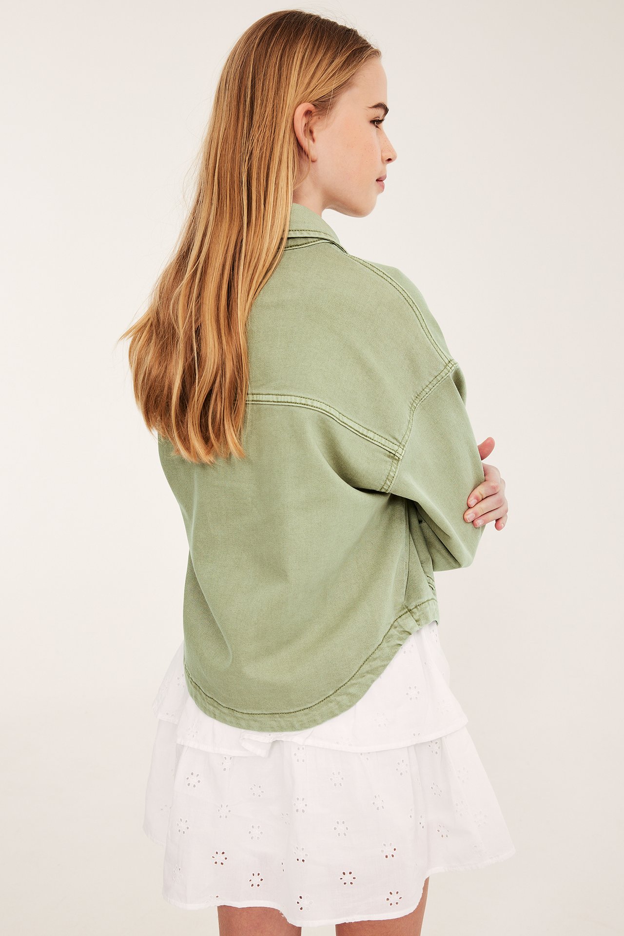 Sidovy av ett barn i en grön jacka av tvättad bomullstwill över en vit broderad kjol.