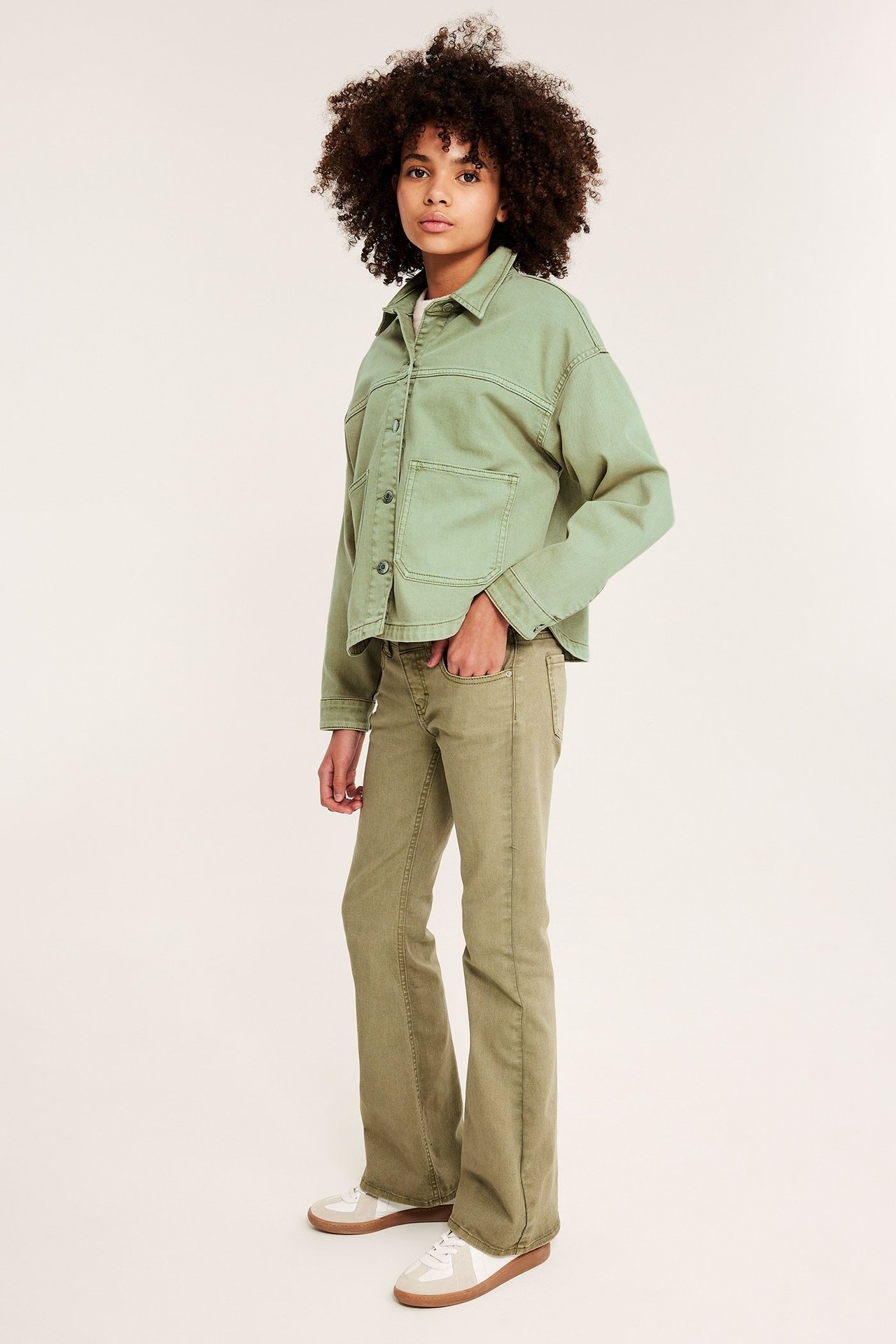 Framifrån, ett barn i grön jacka av tvättad bomullstwill, gröna utsvängda jeans och vita sneakers.