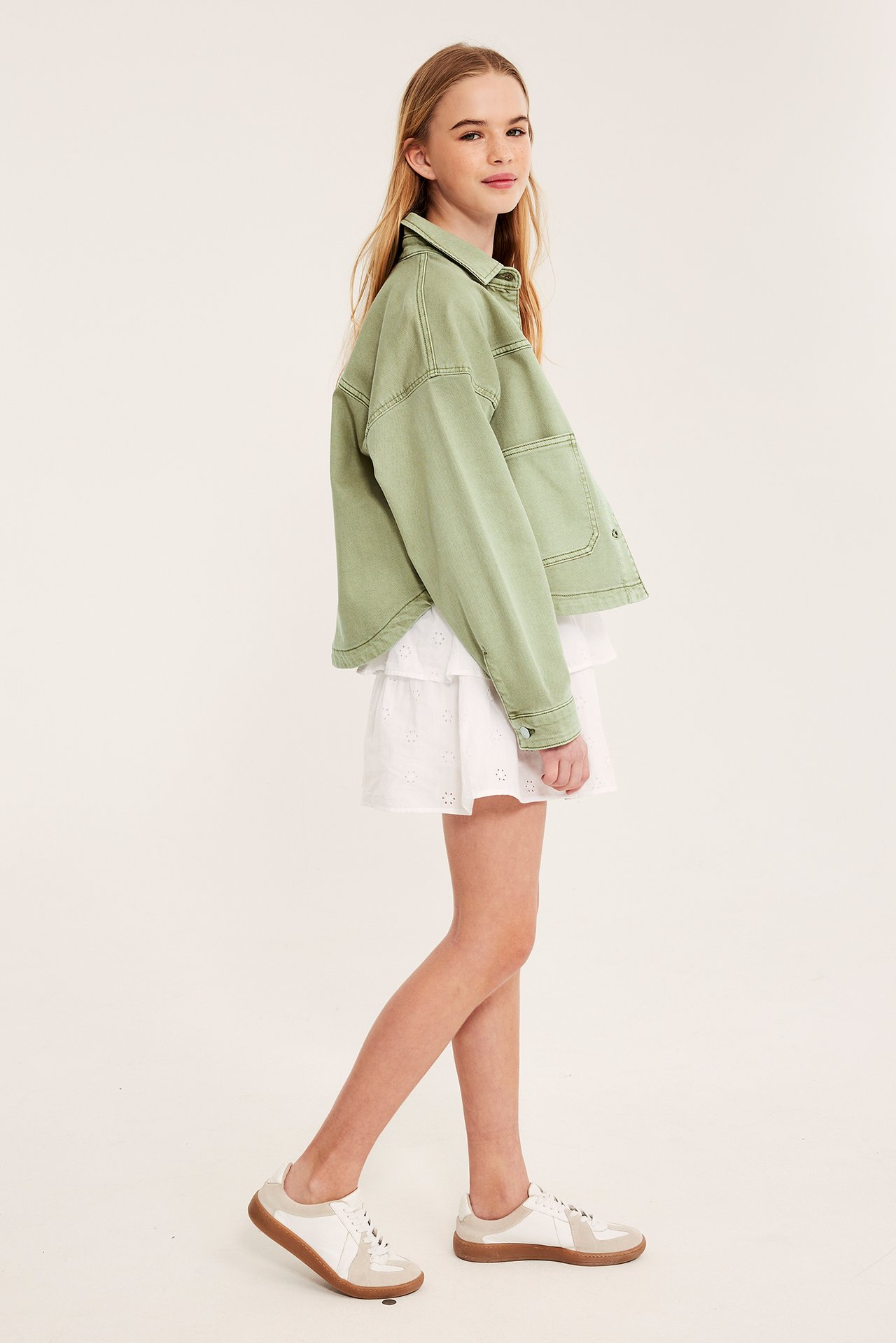 Sidovy av ett barn i en tvättad ljusgrön bomullstwilljacka, vit volangkjol och vita/beige sneakers.