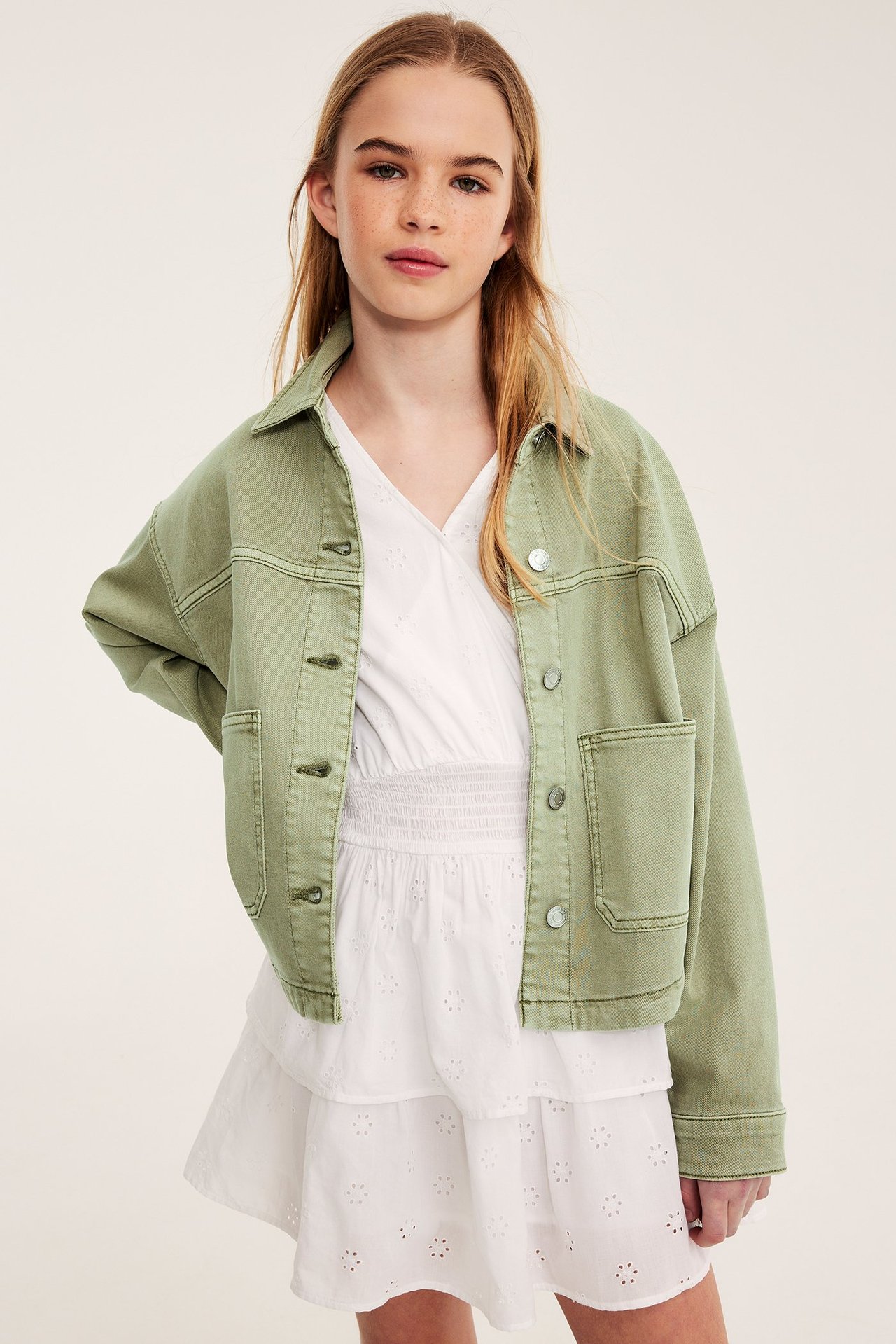 Frontbild av ett barn i en öppen grön tvättad bomullstwilljacka över en vit broderad klänning.