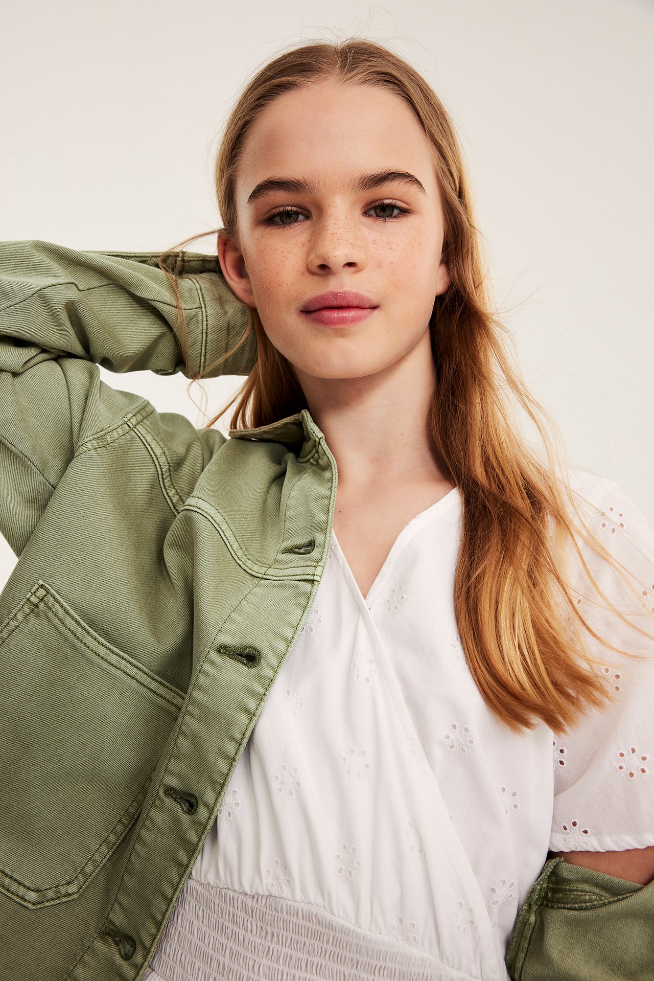 Närbild på ett barn i öppen grön tvättad bomullstwilljacka över en vit topp med broderat mönster.