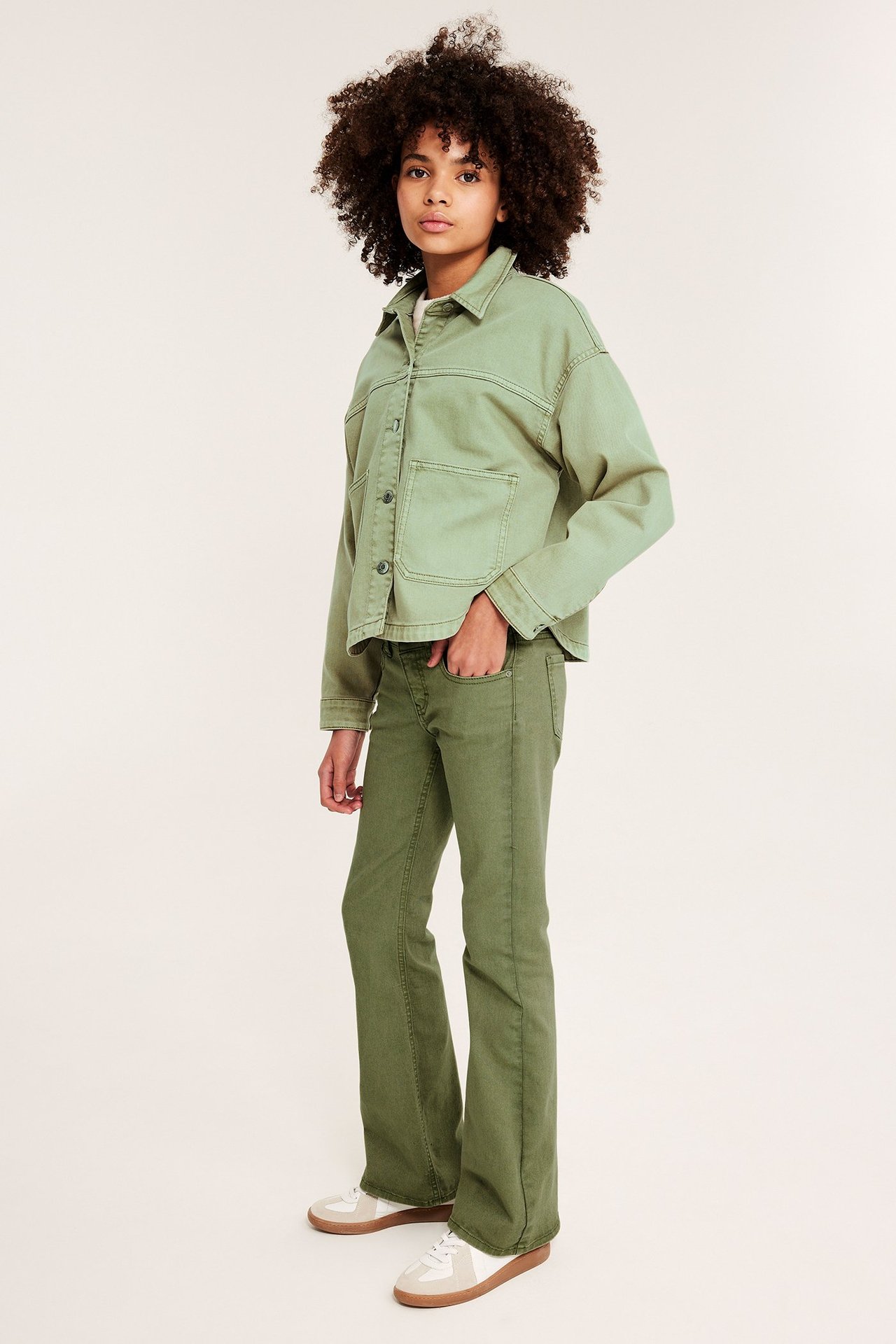 Edestä kuvattu lapsi yllään vihreä pesty puuvillatvillitakki, vihreät levenevät housut ja valkoiset tennarit.