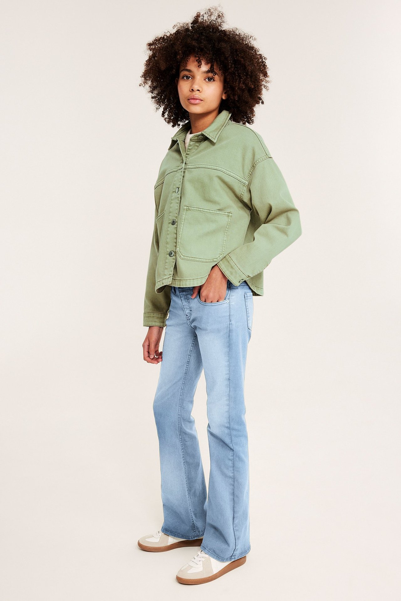 Framifrån, ett barn i grön jacka av tvättad bomullstwill, ljusblå utsvängda jeans och vita/beiga sneakers.