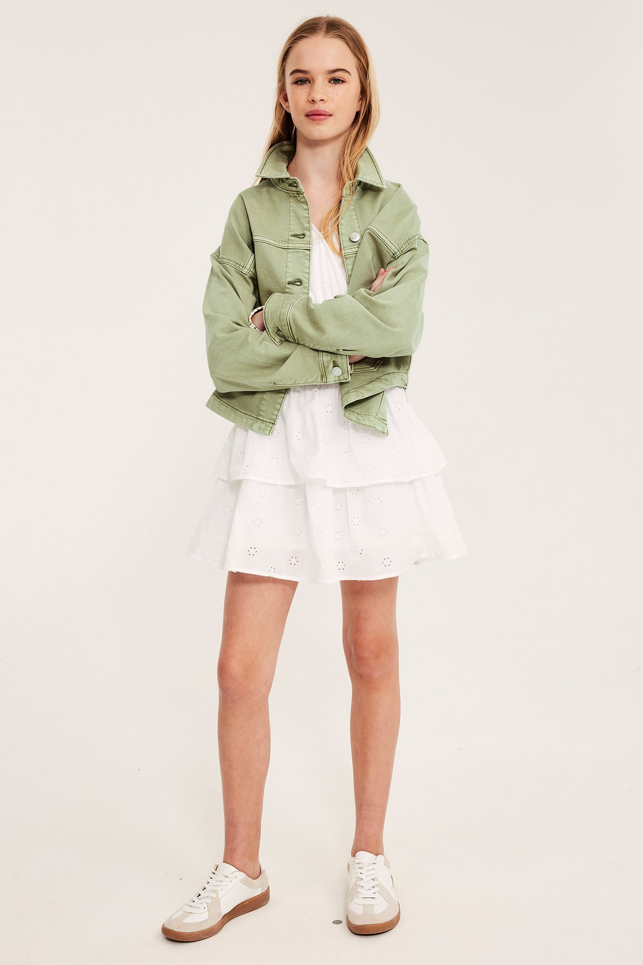 Framifrån: Ett barn i en grön jacka i tvättad bomullstwill över en vit volangklänning och vita sneakers.