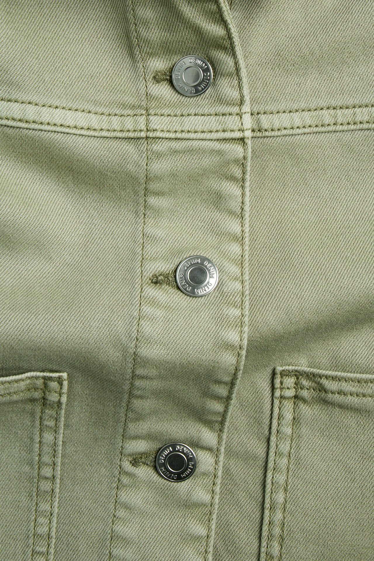 Lähikuva vihreästä pestyä puuvillatvilliä olevasta takista, jossa on hopeanväriset metallinapit ja näkyvät ompeleet.