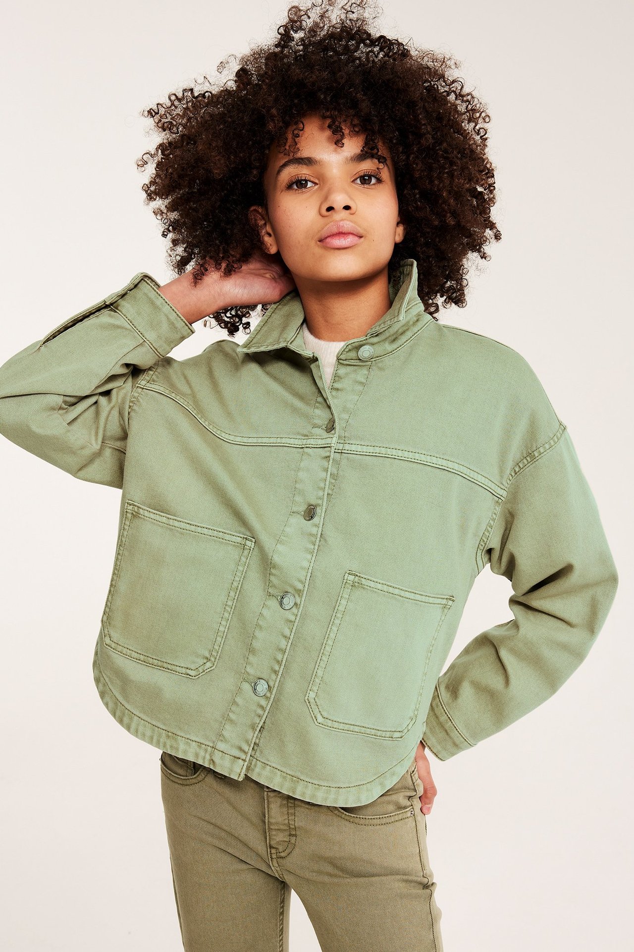 Frontvy: Kid bär en grön tvättad bomullstwilljacka med knappar, patchfickor, ljus topp och gröna byxor.