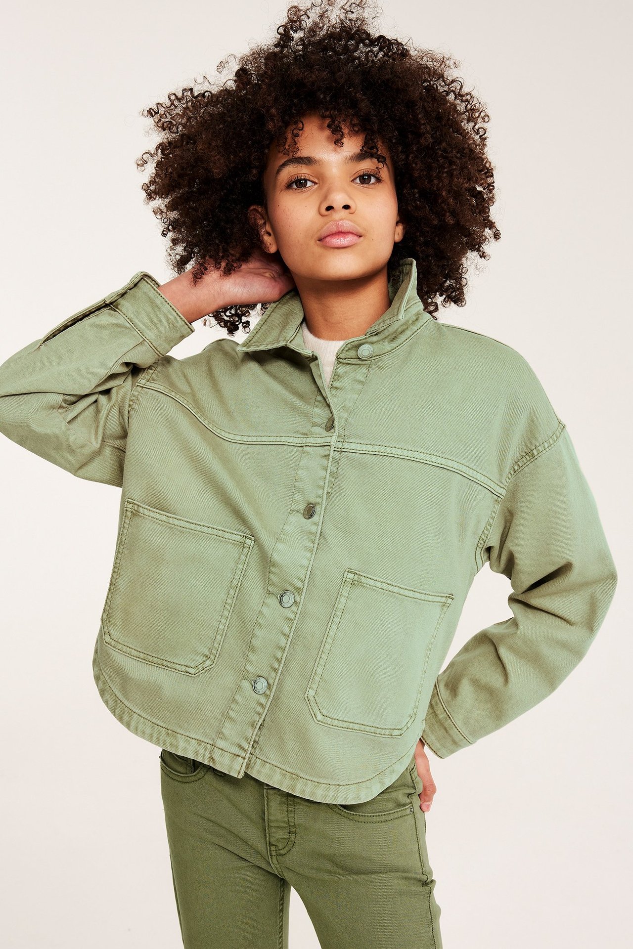 Framifrån: Ett barn bär en grön bomullstwilljacka med knappar och fickor, samt gröna byxor.