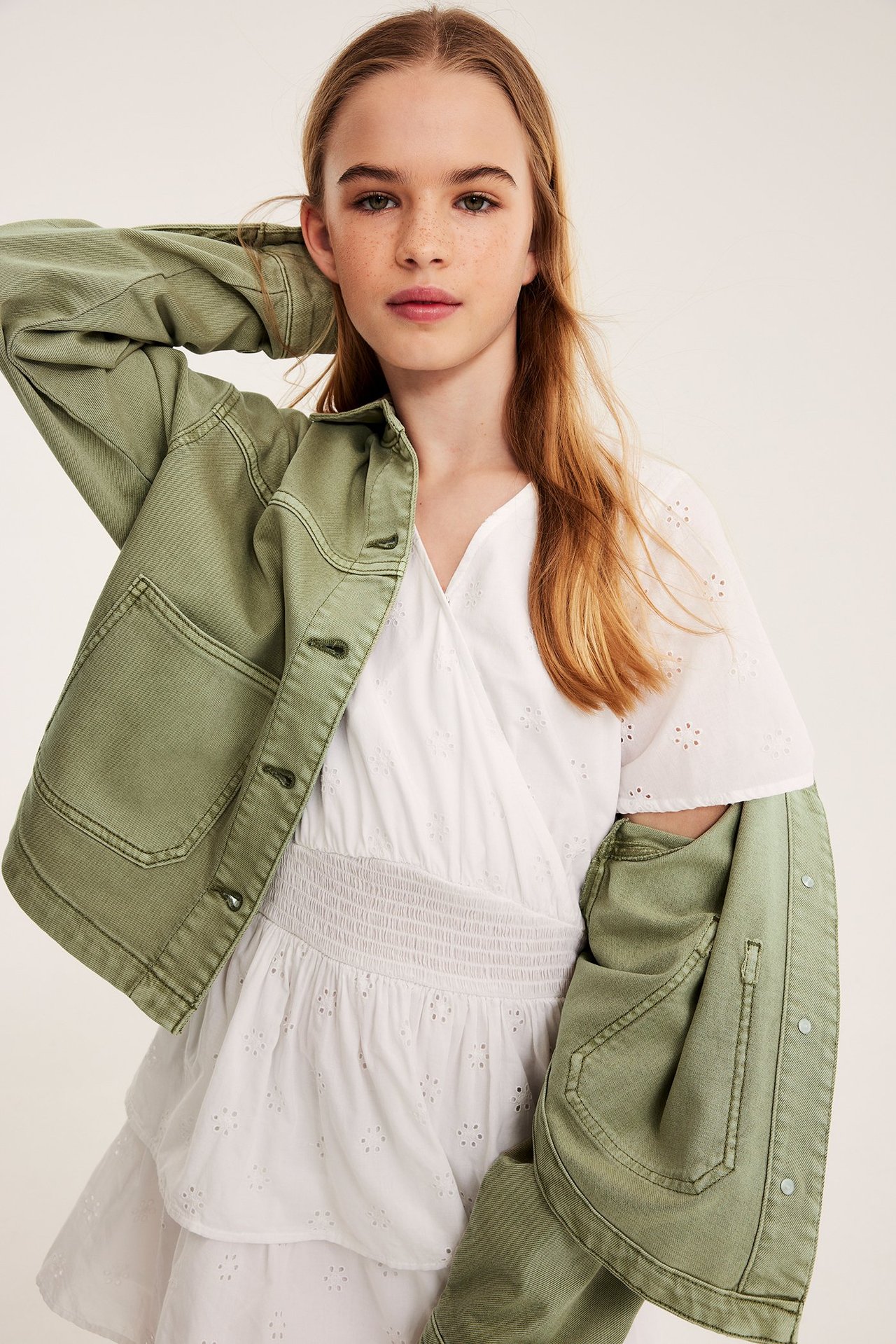 Zbliżenie z przodu: dziecko w zielonej kurtce z dekatowanego twillu bawełnianego na białej sukience z haftem.