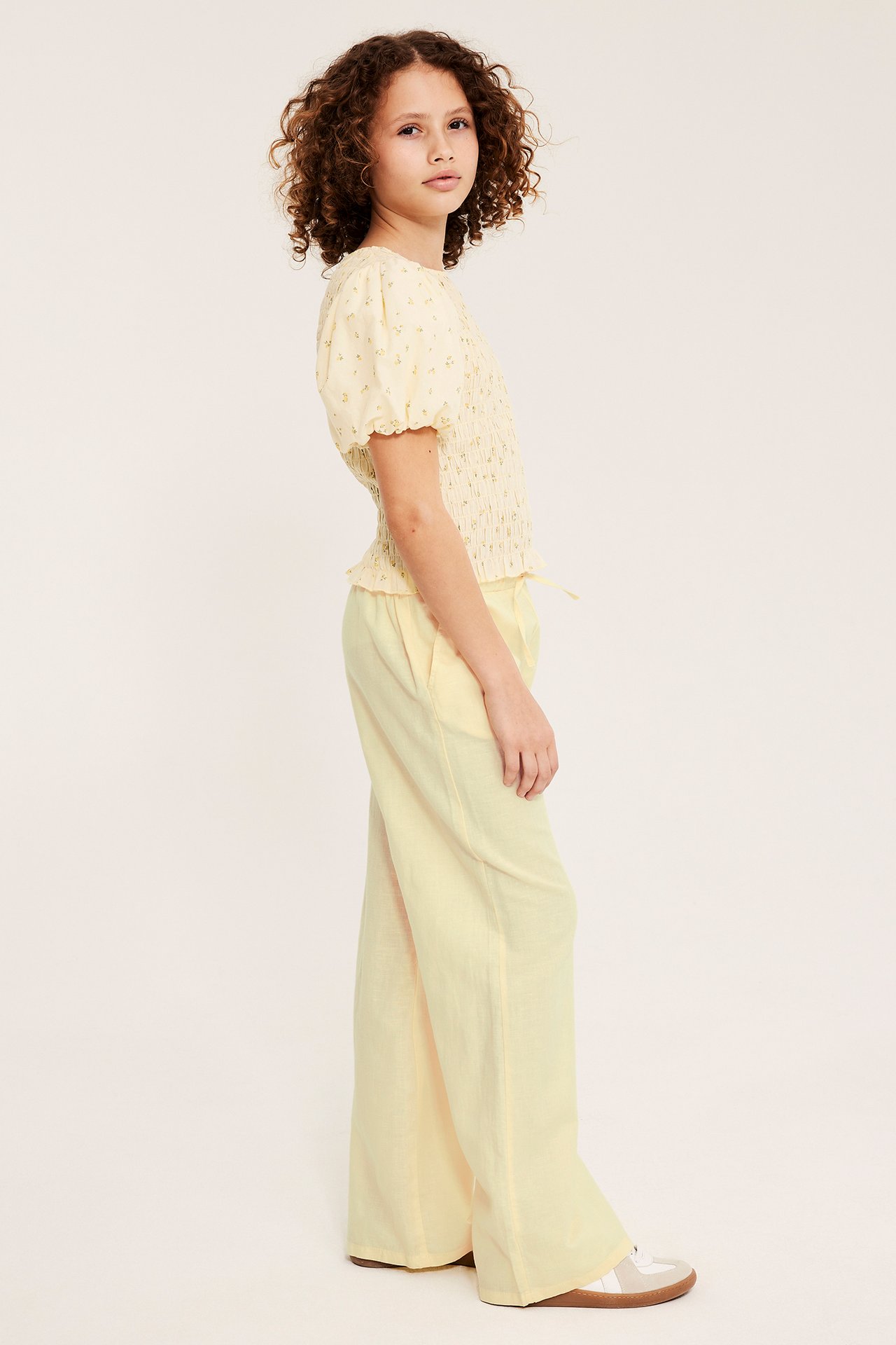 Sidovy: Kid i ljusgula linnebyxor med elastisk dragsko i midjan, blommig topp och sneakers.