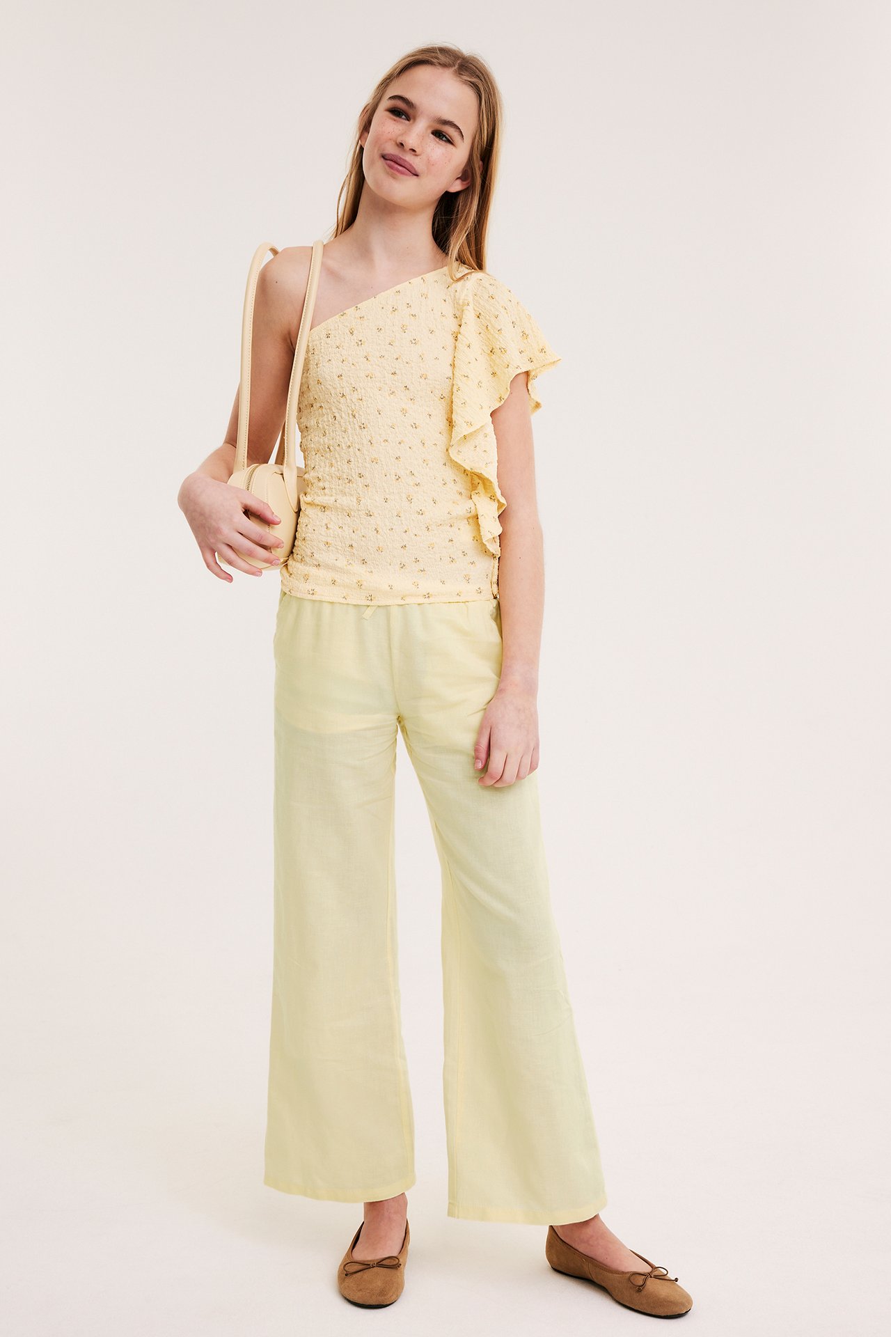 Barn i ljusgula linnebyxor med vida ben, blommig enaxlad topp, bär beige väska. Framifrån.