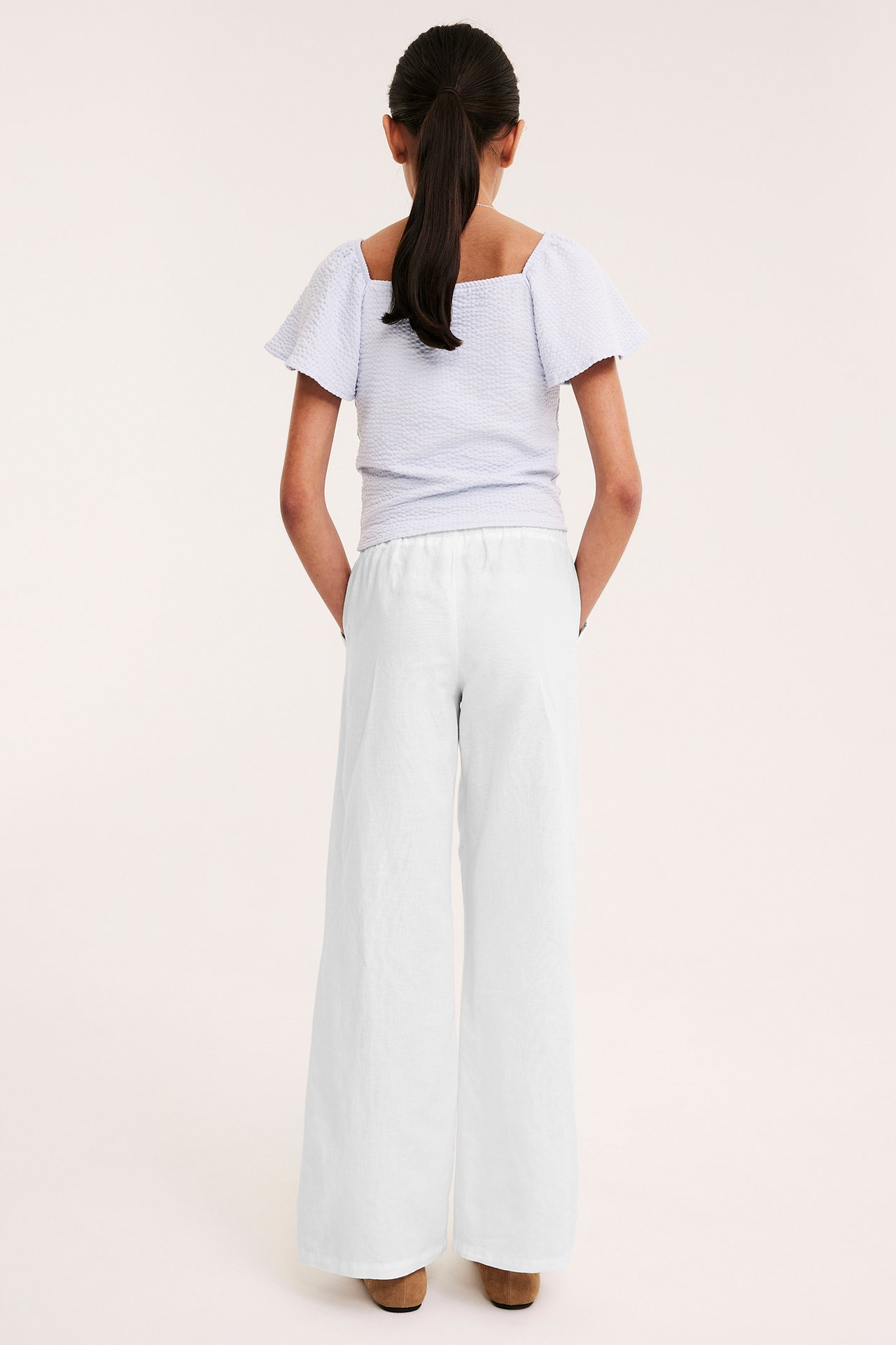 Ryggvy av ett barn i vita, vida linnebyxor med resår i midjan och en ljusblå texturerad topp.