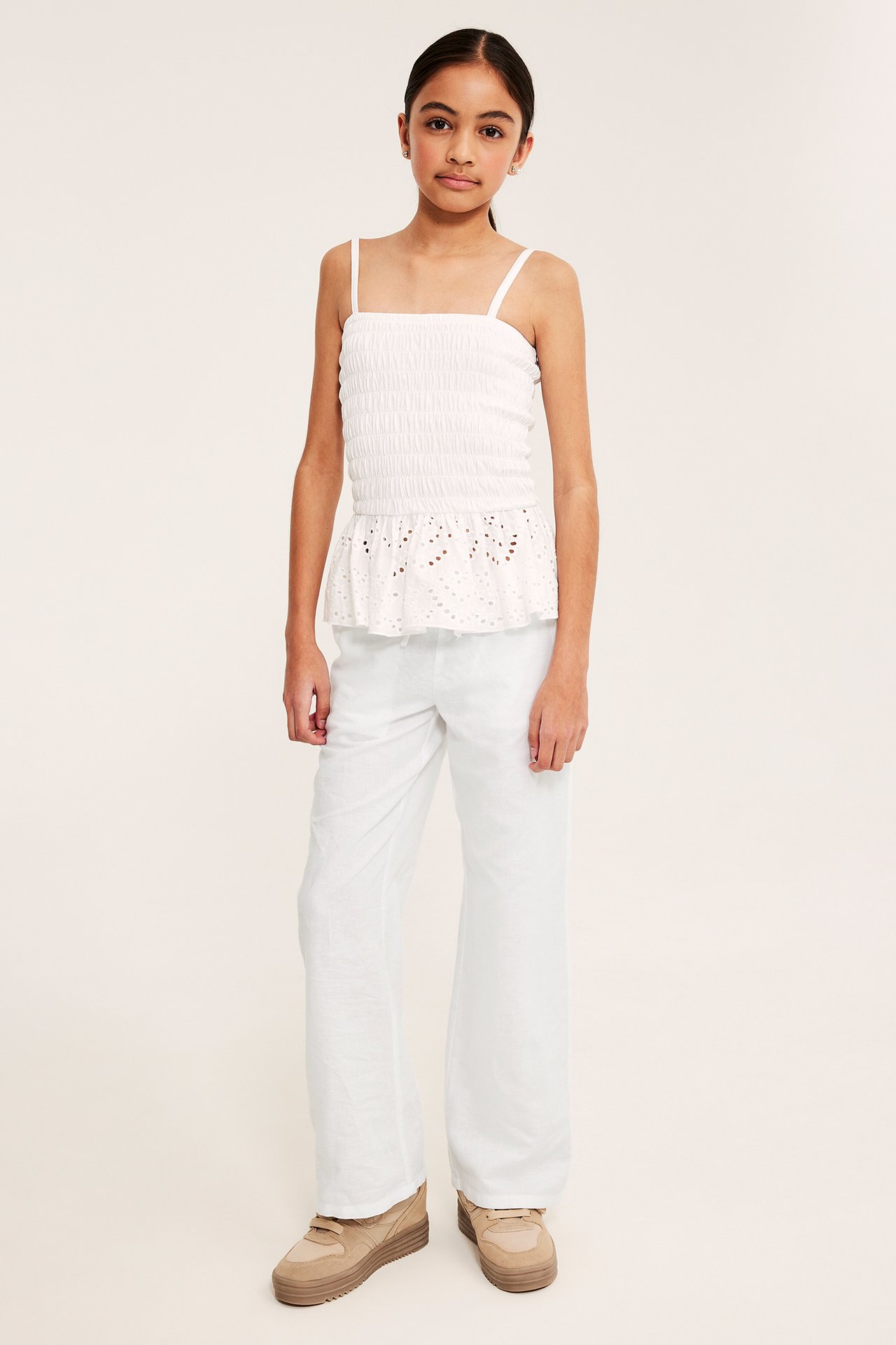 Przód: dziecko w białych, szerokich spodniach z lnu, białej marszczonej koszulce i beżowych trampkach.