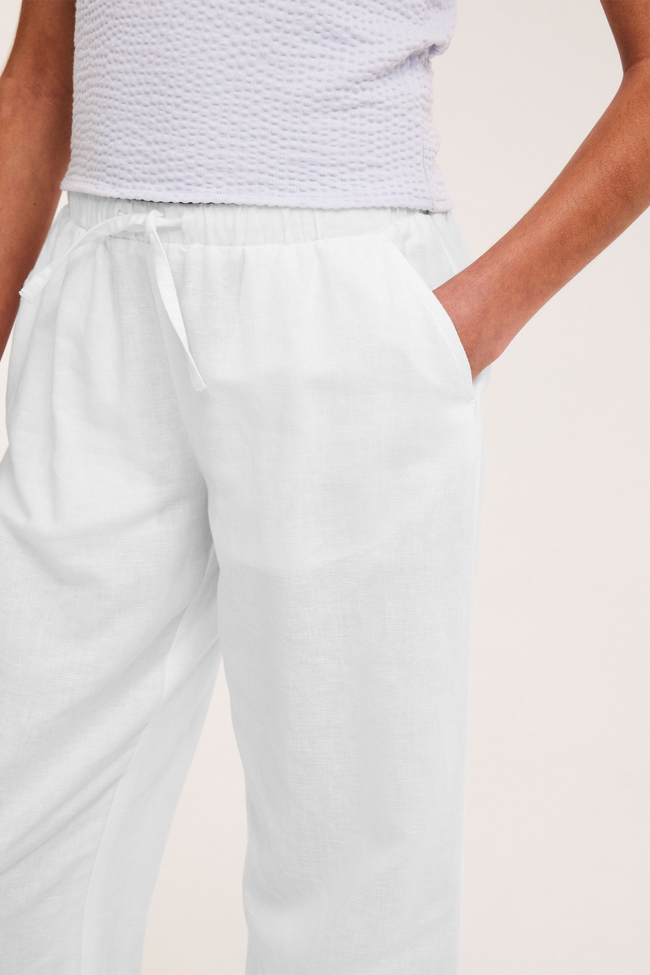 Närbild: kvinna i vita linneblandningsbyxor med dragsko, hand i fickan, och ljusblå topp.