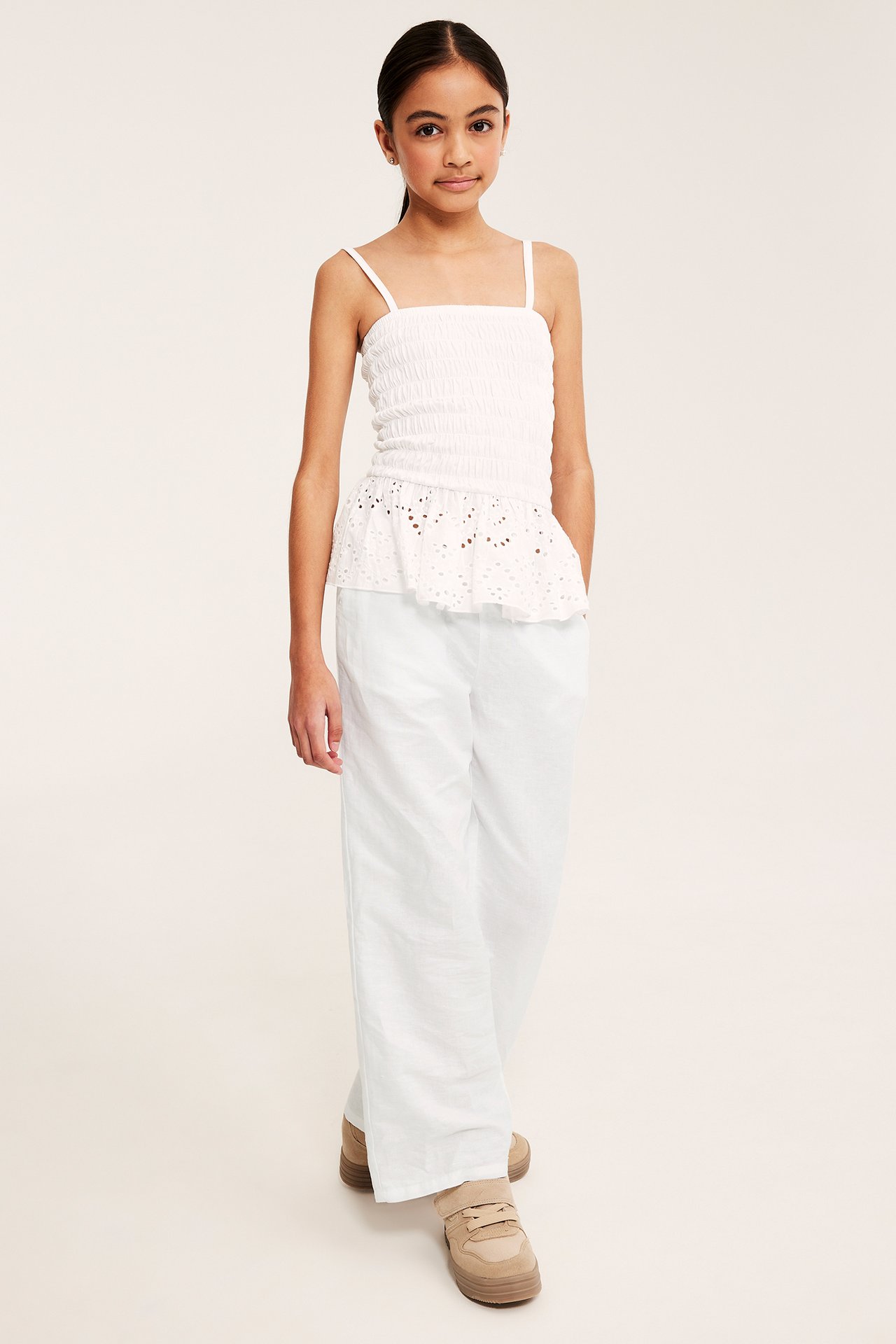 Widok z przodu: dziecko w białych szerokich spodniach z lnu, białym marszczonym topie i beżowych sneakersach.