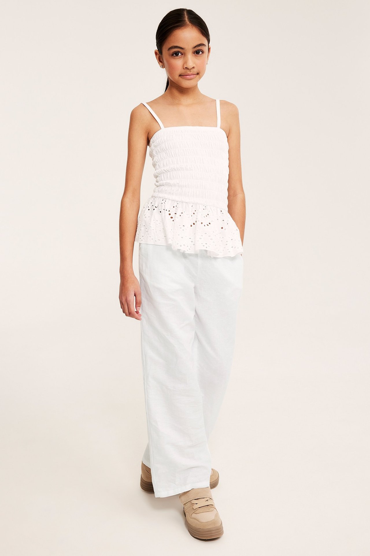 Frontvisning av et barn i vide hvite linblandingbukser, en hvit smocketopp og beige joggesko.