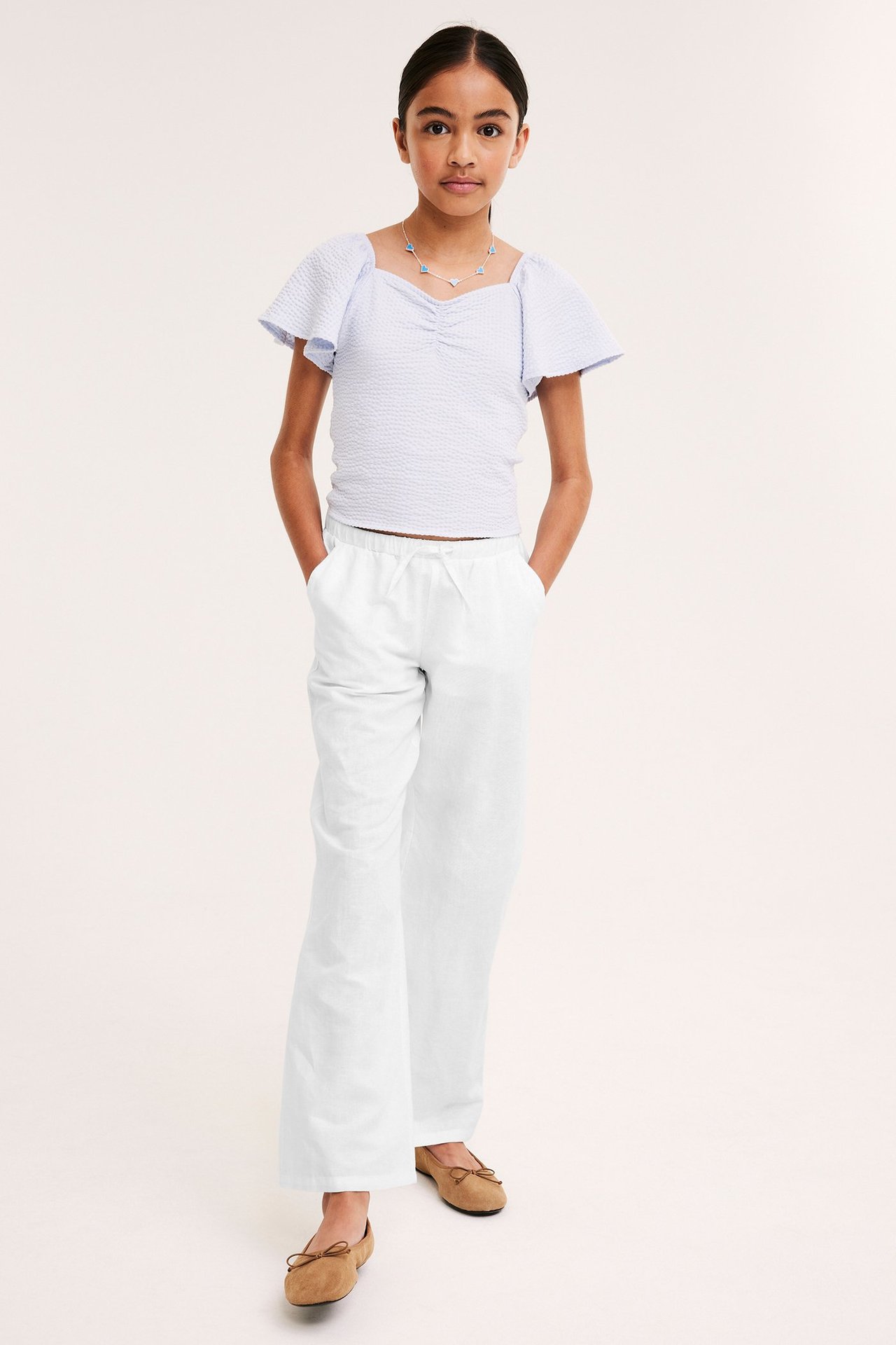 Etunäkymä lapsesta valkoisissa pellavahousuissa nyöreillä, vaaleansinisessä topissa ja ruskeissa ballerinat.