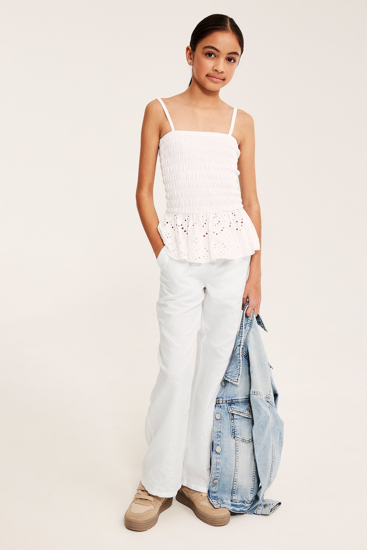 Barn bär vita linneblandningsbyxor, en vit smockad topp, och håller en ljusblå jeansjacka.