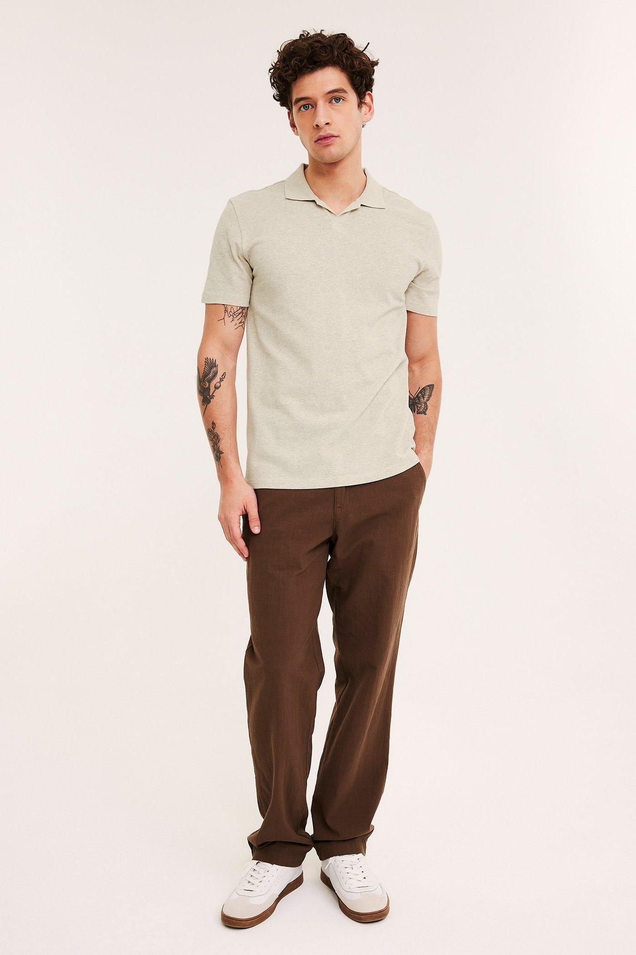 Edestä, mies yllään beigemelange pikeepaita, ruskeat housut ja valkoiset tennarit.