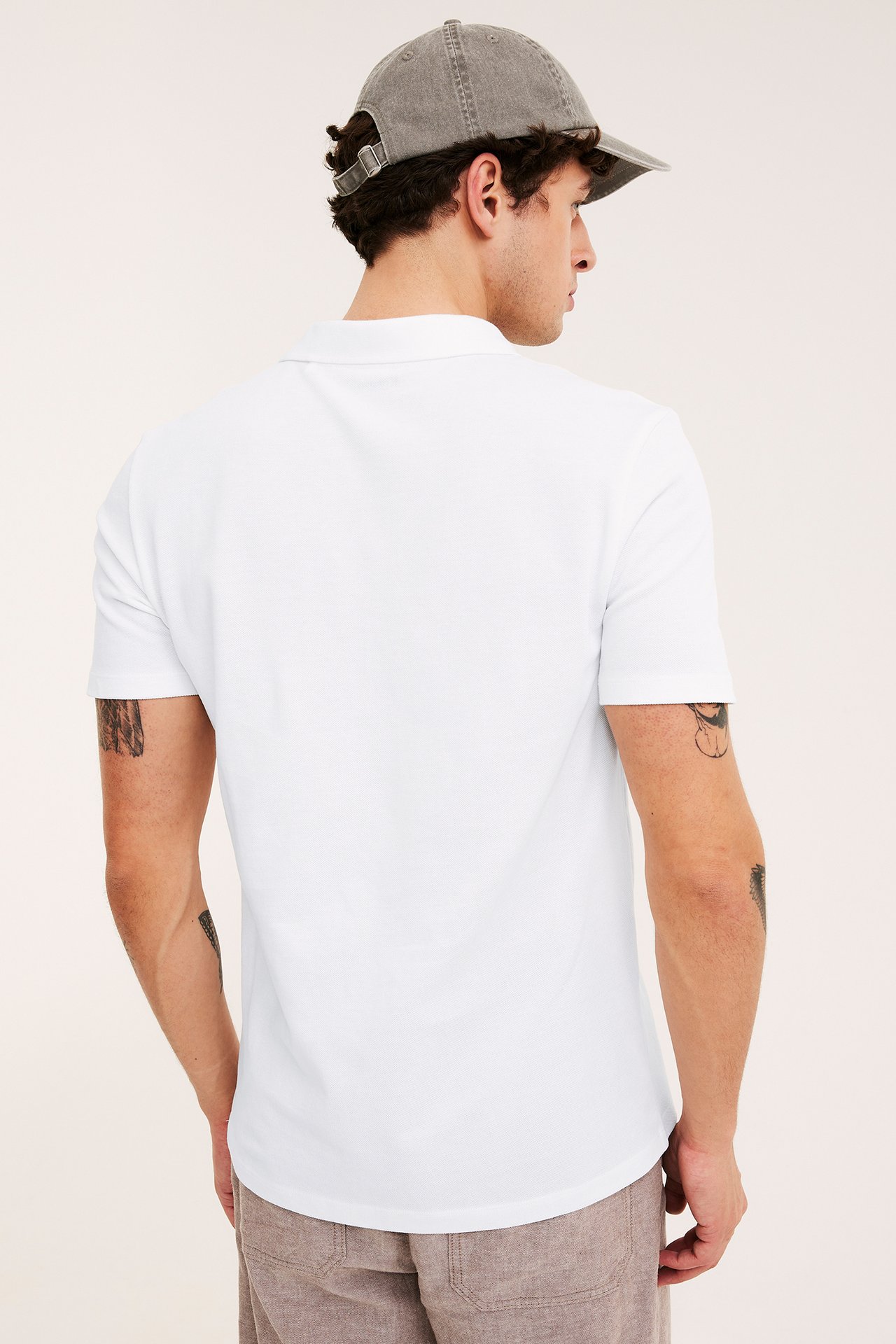 Bakfra/sidevisning av en mann i en hvit bomullspikétrøye, en grå caps og lysebrune shorts.