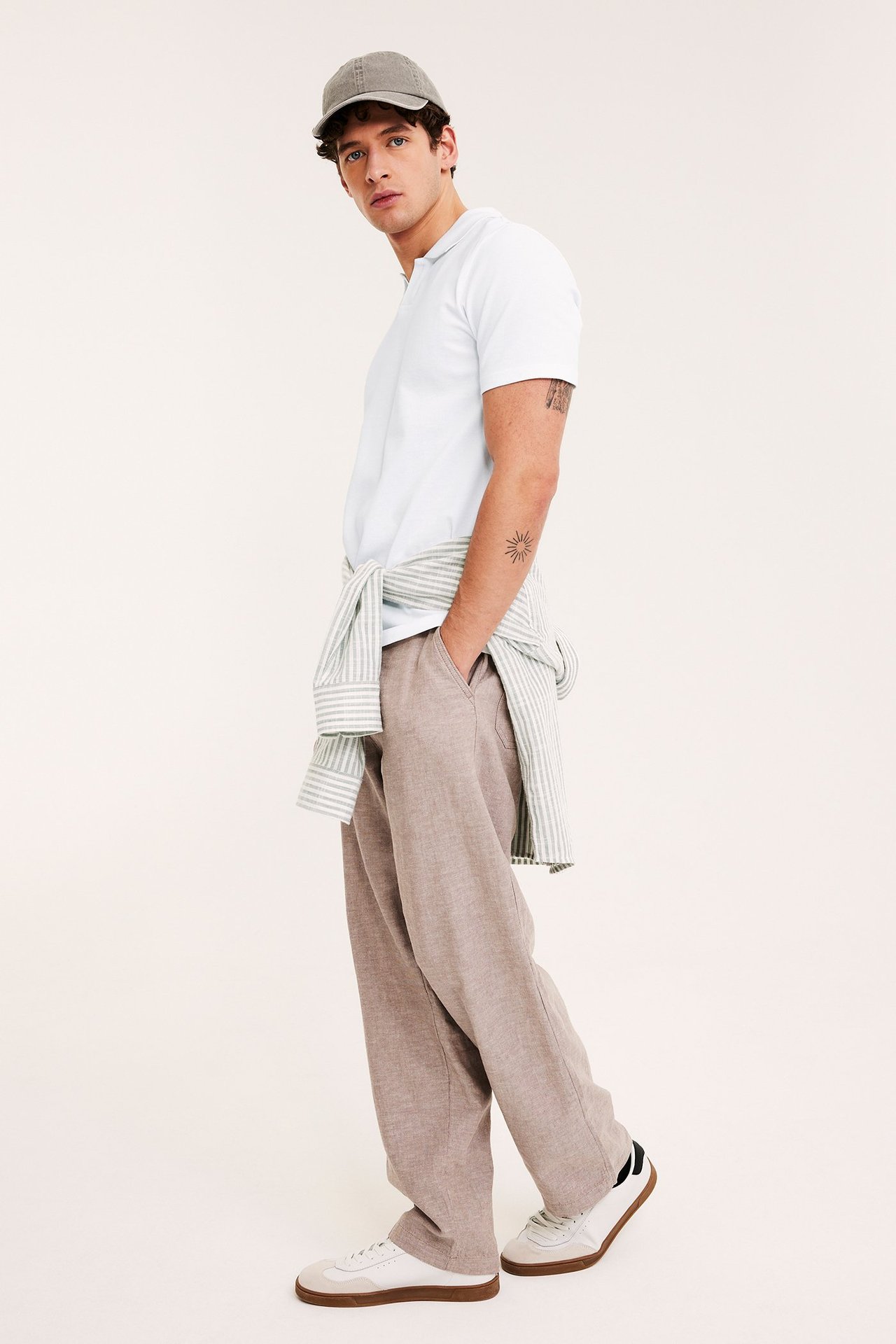 Mies edestäpäin yllään valkoinen pikeepaita, vaaleanruskeat housut ja harmaa lippalakki.