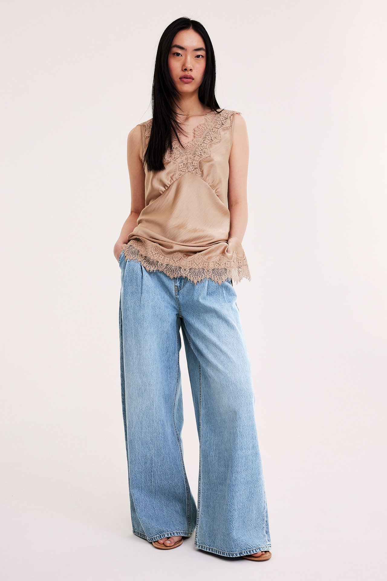 Nainen edestäpäin, yllään vaaleansiniset leveälahkeiset korkeavyötäröiset farkut ja beigenvärinen pitsikoristeltu hihaton toppi.