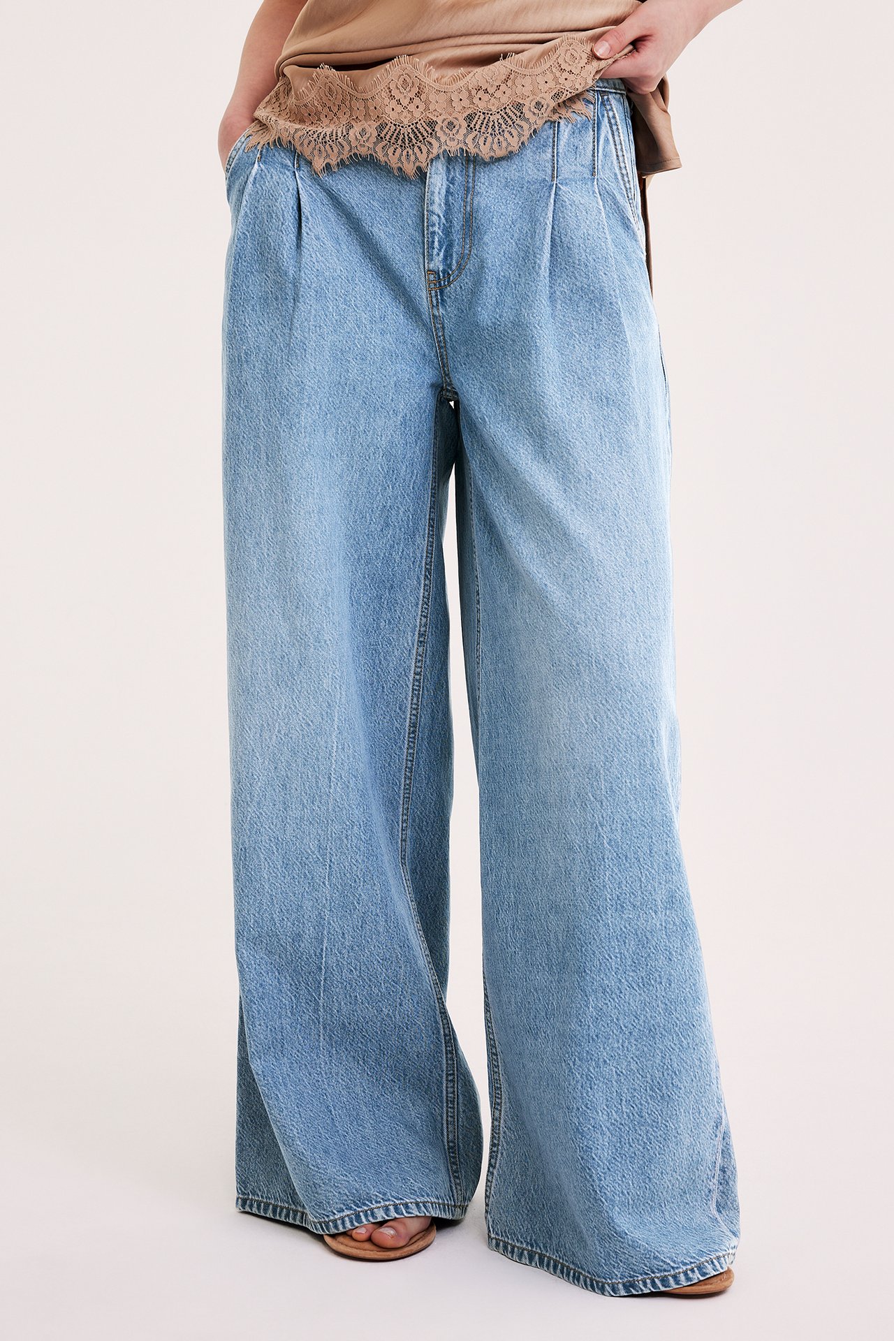 Nainen vaaleansinisissä leveälahkeisissa farkuissa laskoksilla, beige pitsitoppi, sandaalit.