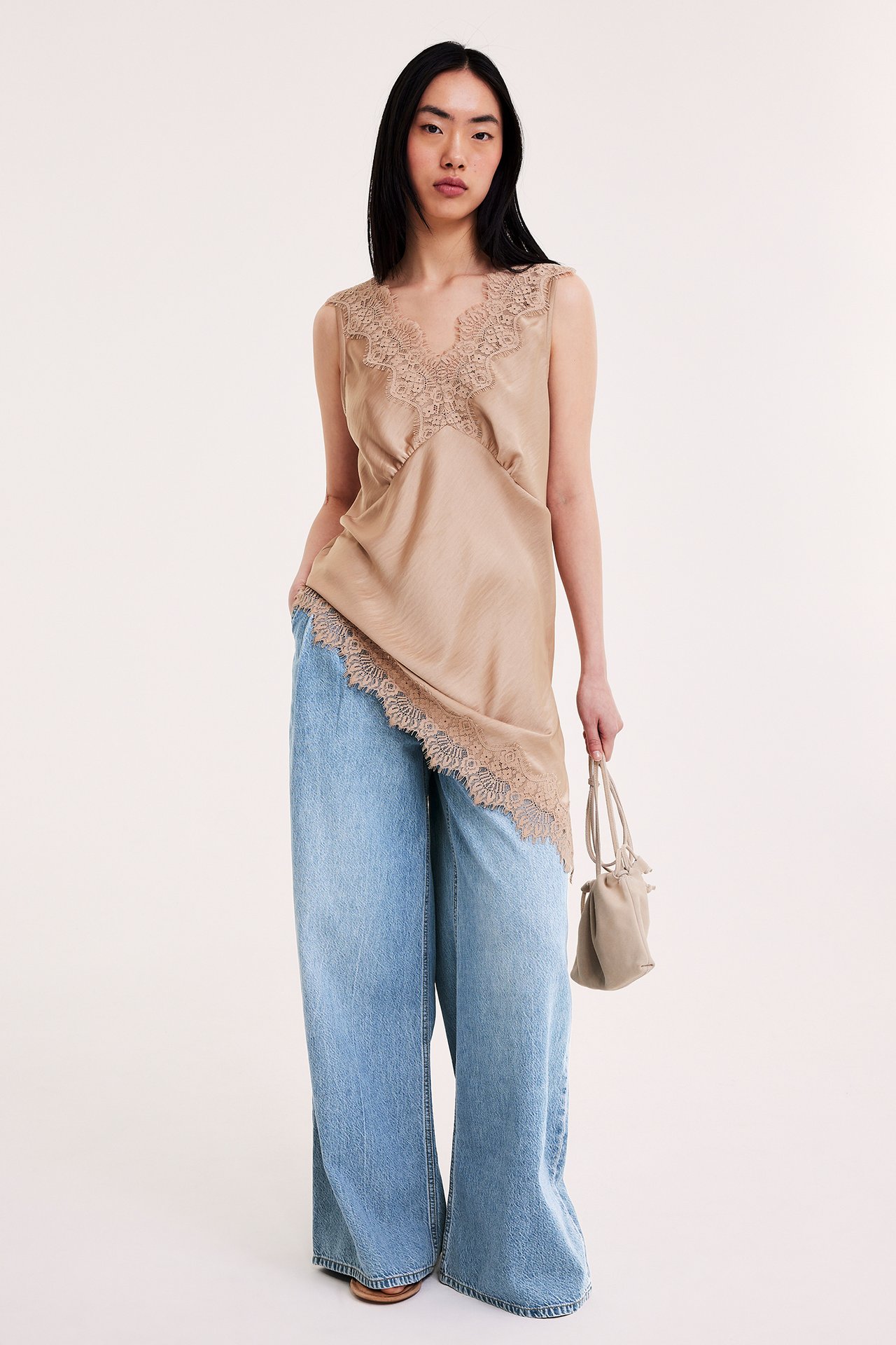 Edestäpäin: Nainen vaaleissa leveissä farkuissa laskoksilla, beigessä pitsitopissa, pitäen pientä beigeä laukkua.