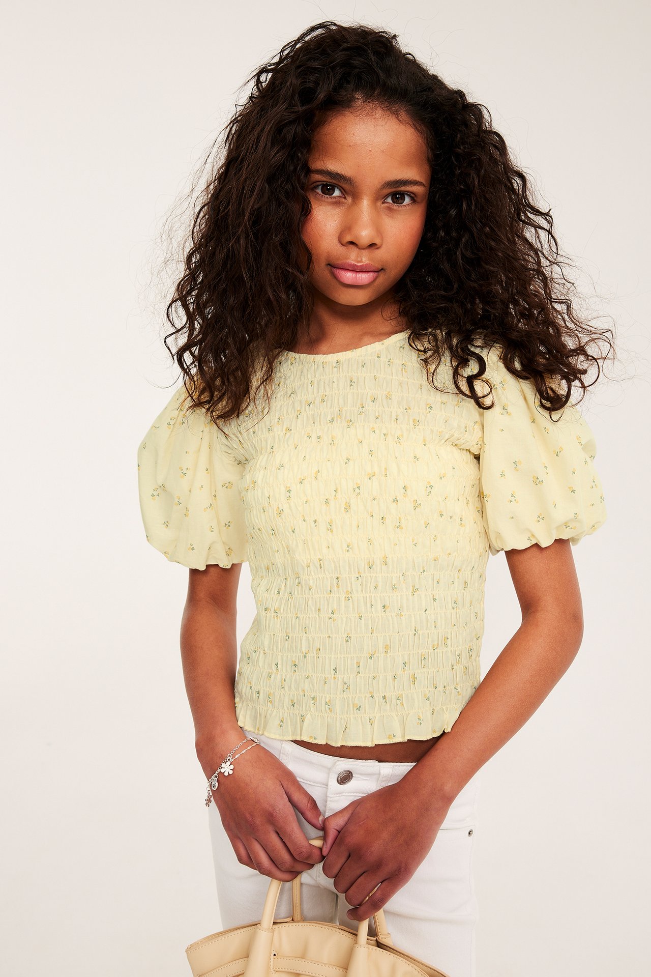 Barn har på seg en lysegul blomstret smocktopp med puffermer, hvite jeans, sølvarmbånd, holder beige veske.