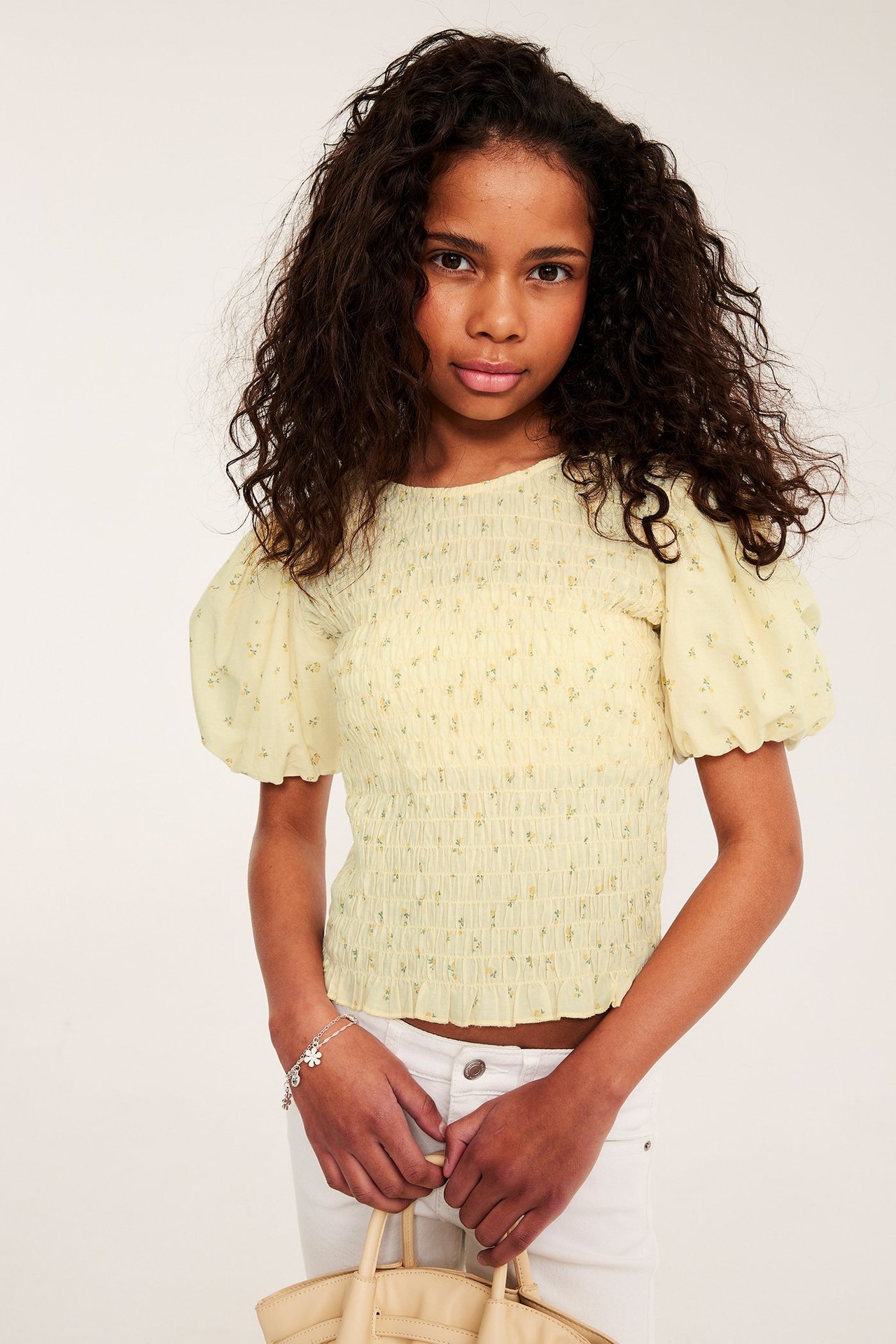 Barn bär en ljusgul blommig smocktopp med puffärm, vita jeans, silverarmband, håller beige handväska.