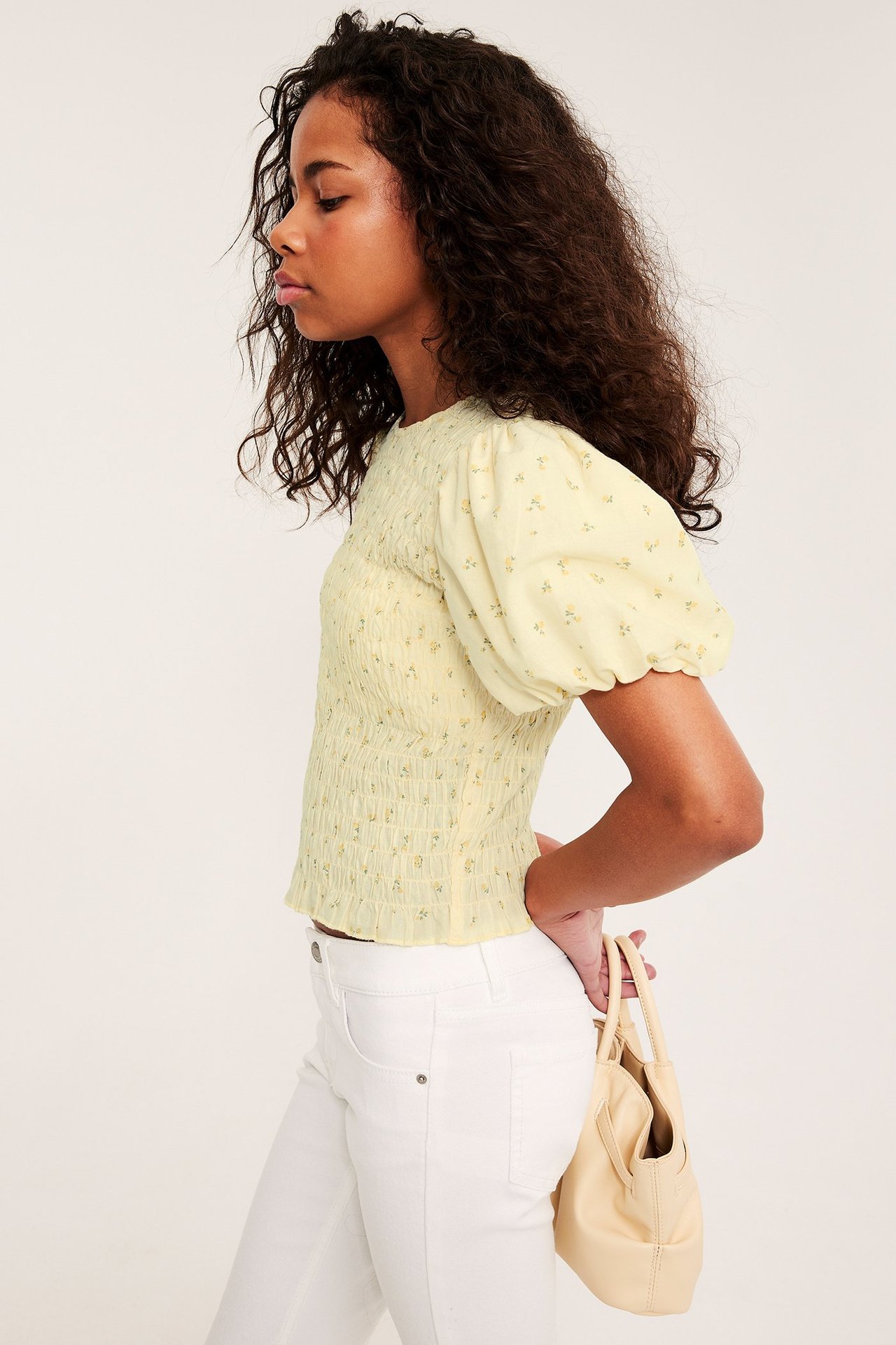 Sivukuva lapsesta vaaleankeltaisessa kukkakuvioisessa smokkitopissa, valkoisissa farkuissa, beigen laukun kanssa.