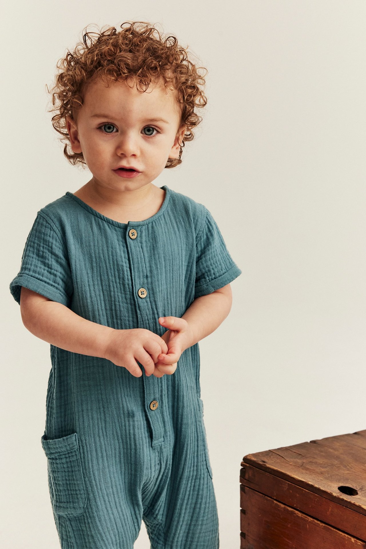Frontvisning av baby i mørk turkis bomullsmuslin jumpsuit med knapper foran og lappelommer.