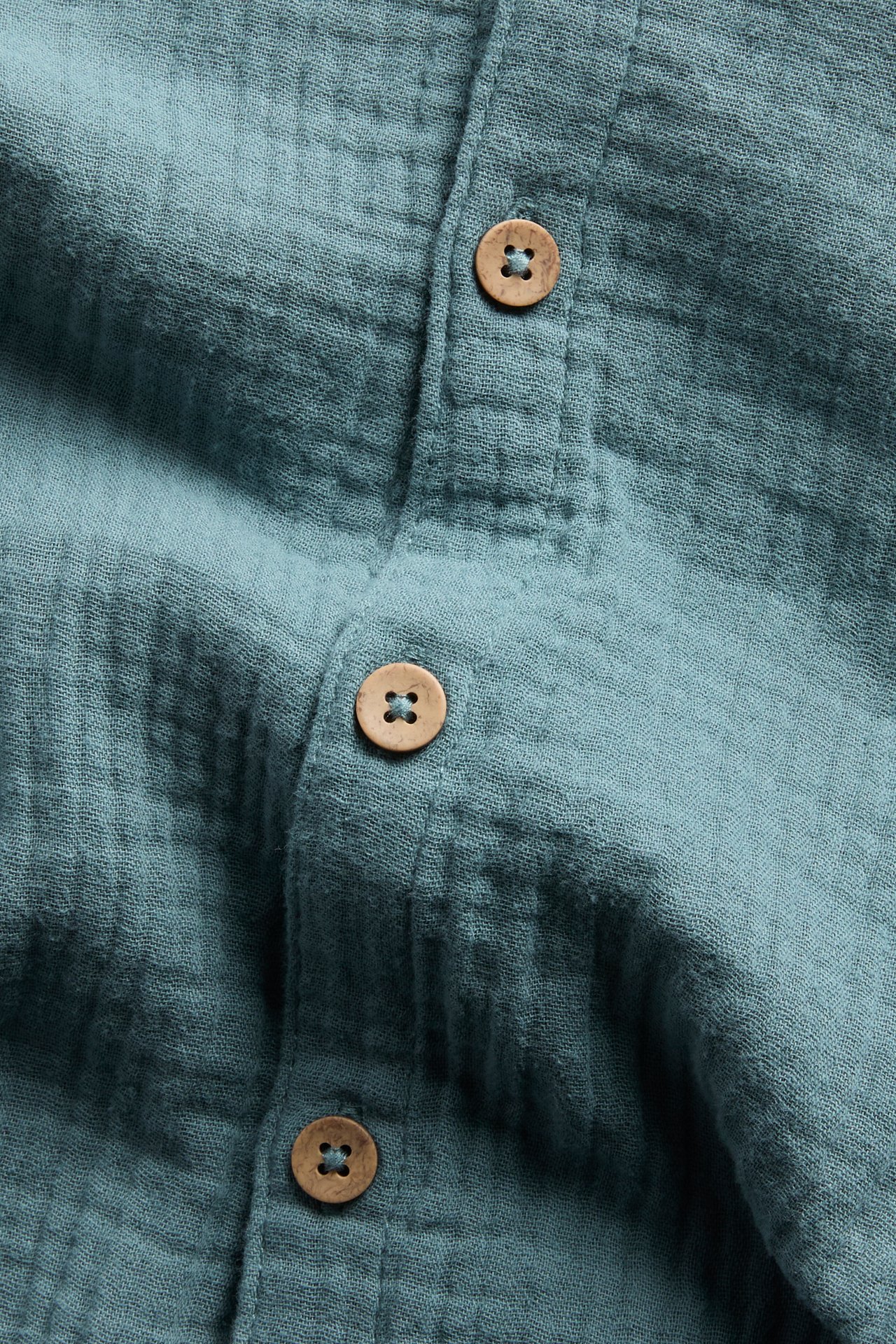 Zbliżenie na ciemnoturkusowy, krótki kombinezon z muślinu z naturalnymi drewnianymi guzikami.