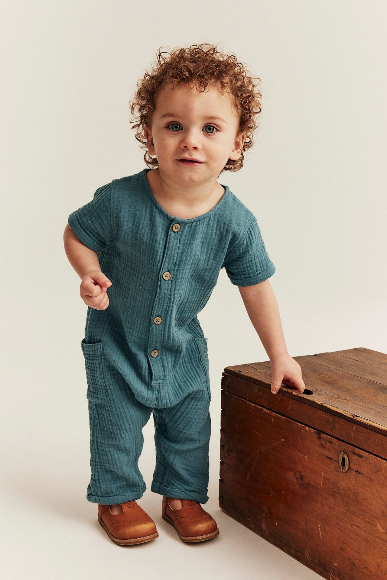 Niemowlę nosi z przodu widoczny, ciemnoturkusowy bawełniany kombinezon z muślinu z guzikami, kieszeniami i brązowymi butami.