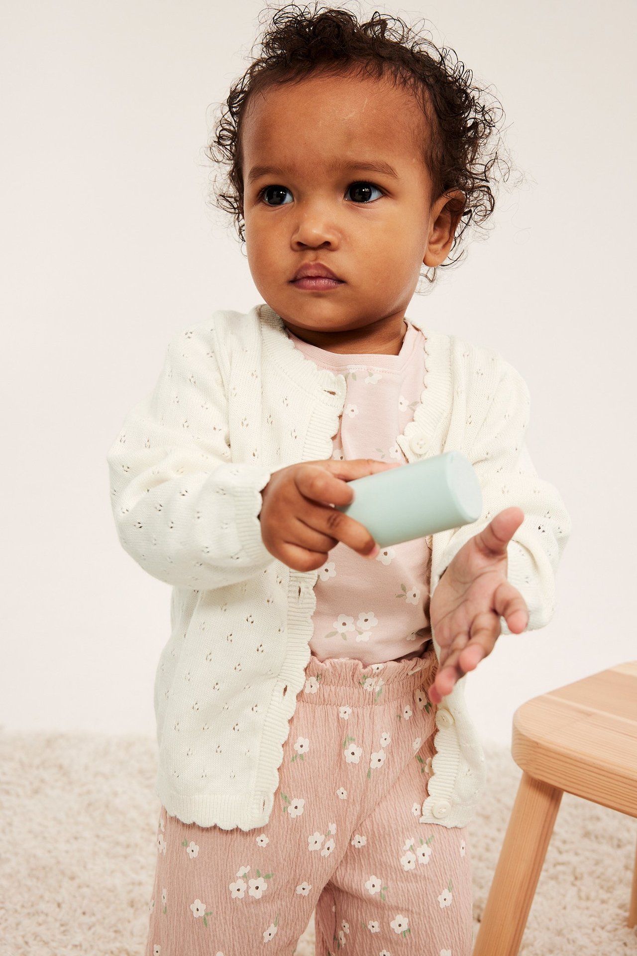 Edestä näkymä vauvasta: luonnonvalkoinen puuvillainen pointelle-neuletakki, vaaleanpunainen kukkatoppi ja housut, lelu kädessä.