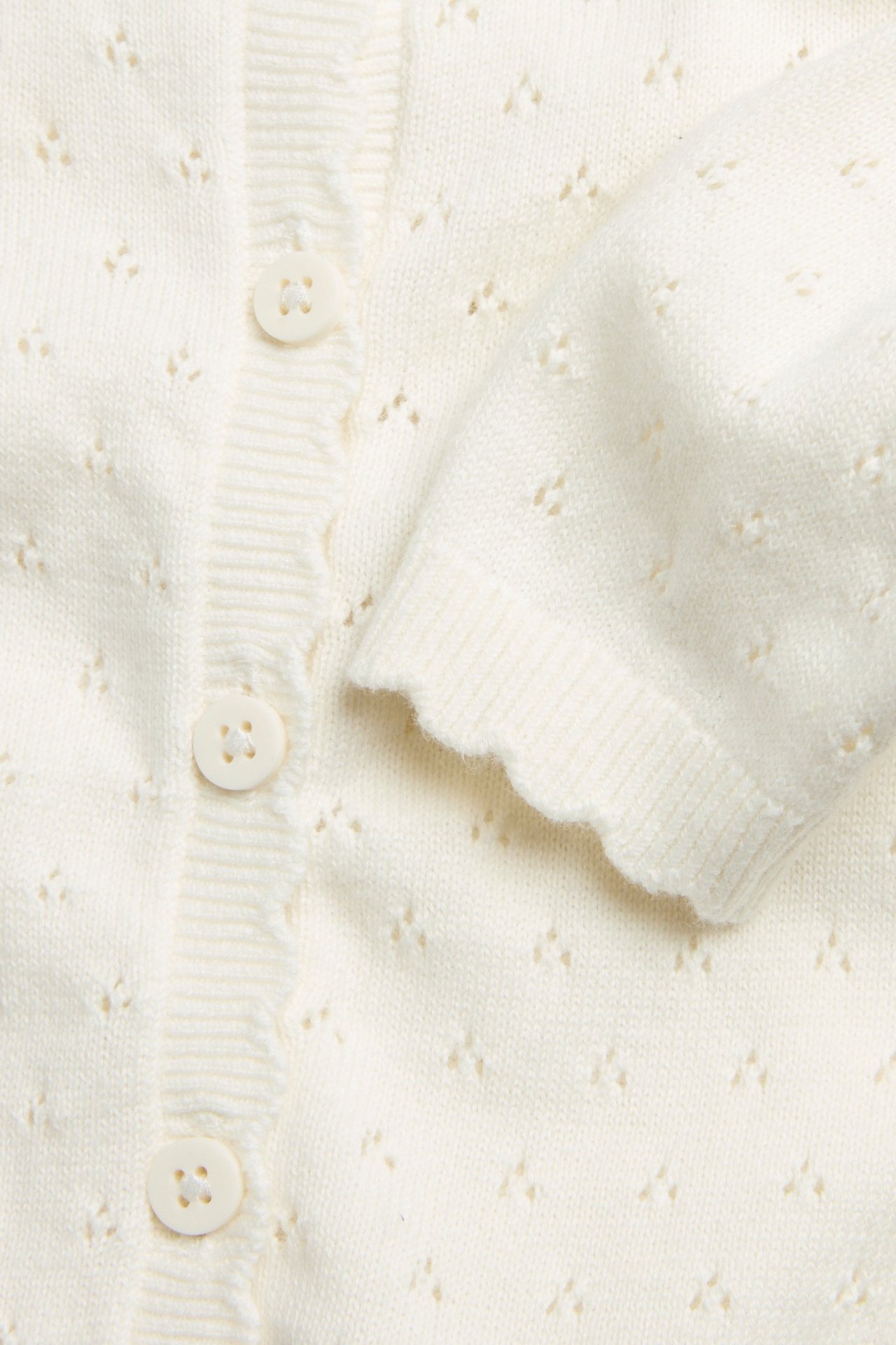 Zbliżenie na kremowy bawełniany sweter rozpinany z wzorem ażurowym, zapięciem na guziki i falowanymi mankietami.