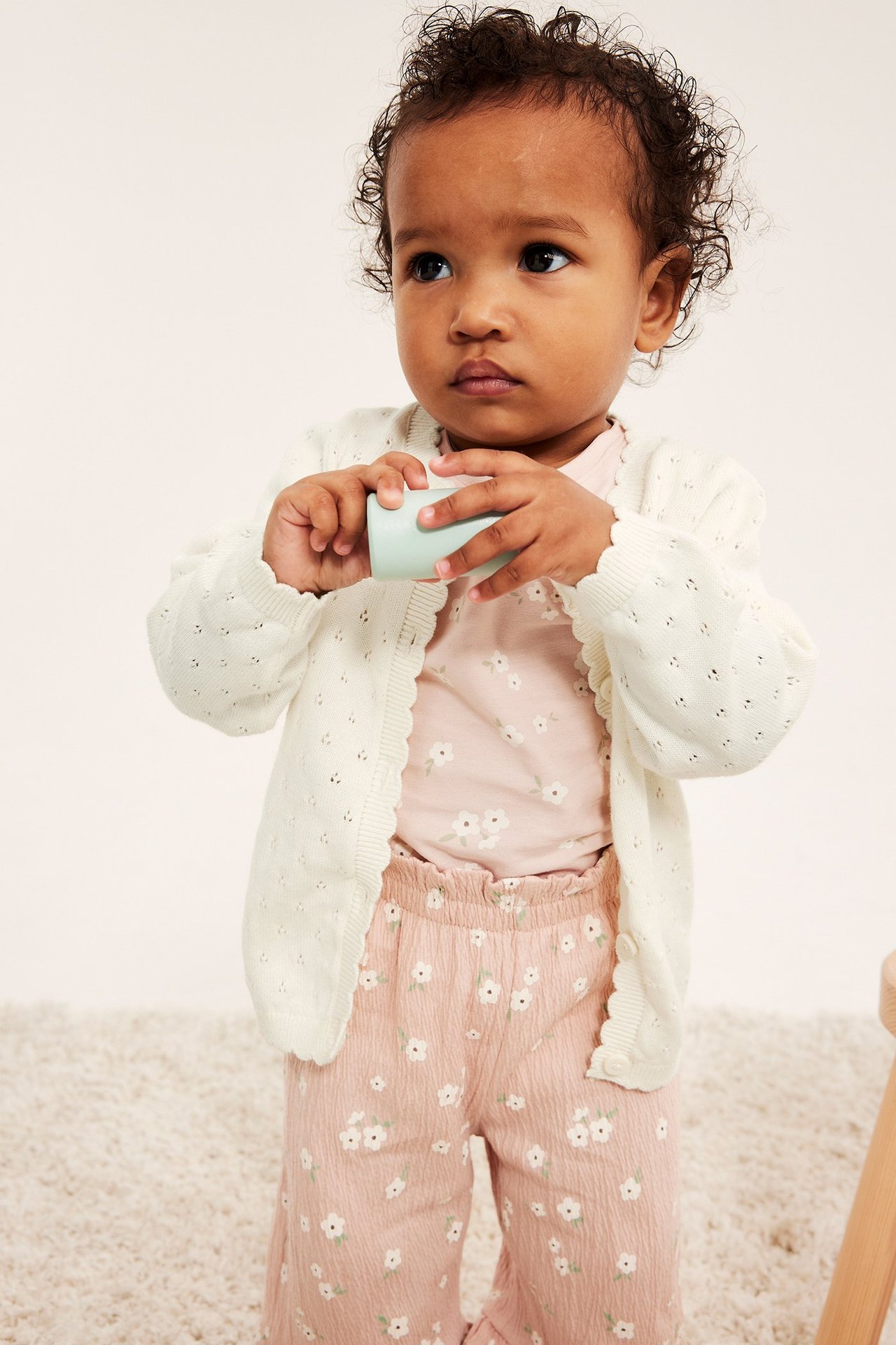 Edestä: Vauva offwhite pointelle-neuletakissa, alla pinkki kukkatoppi ja housut, pitää sinistä mukia.