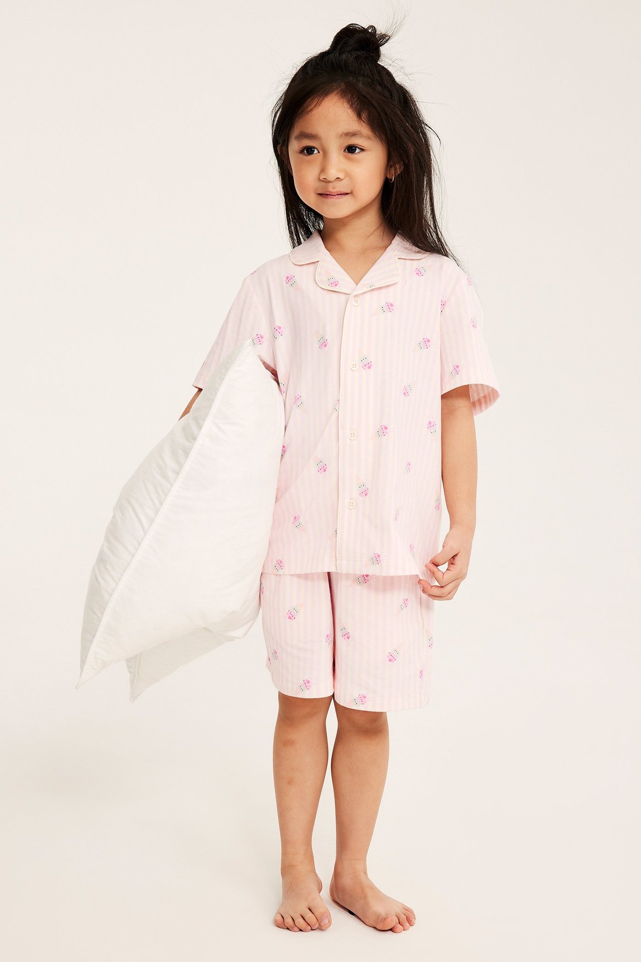 Edestä kuvattuna lapsi pinkkiraidallisessa puuvillapyjamassa jäätelökuviolla, valkoinen tyyny kädessä.